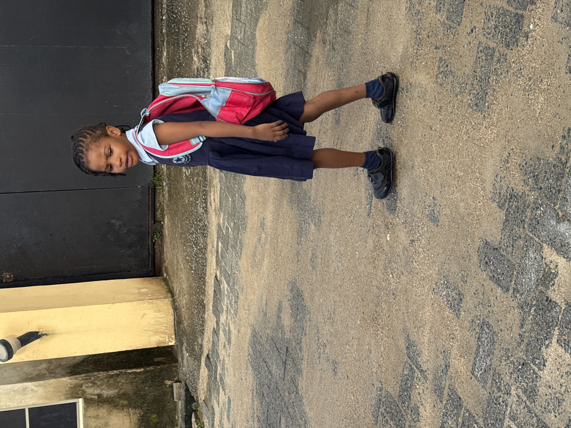 A young Black girl wearing a navy blue school uniform with a light blue collar and a red and blue backpack walks on a paved street in Ado, Nigeria. The pavement is a mixture of sand and interlocking blocks. To her left, a cream-colored building with a dark-colored door is visible. The girl's hair is braided, and she is wearing black velcro shoes and dark blue socks. The scene appears to be outdoors during daylight.