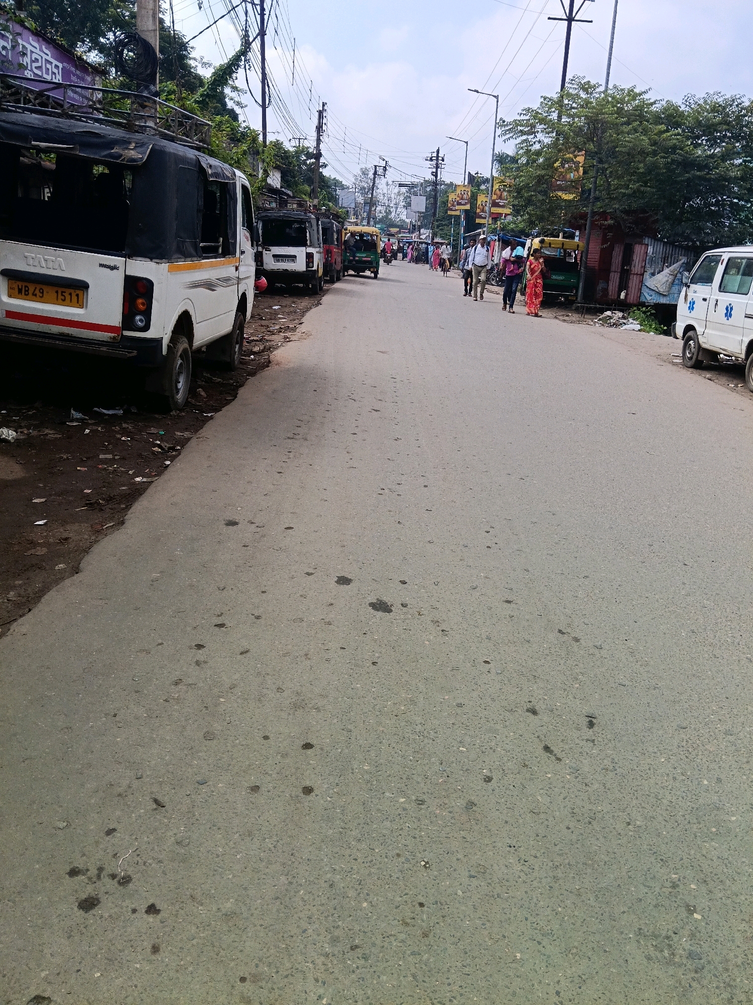 A clear daytime shot captures a street scene in Purulia, India, featuring an assortment of vehicles and people. The foreground is dominated by the rear of a white Tata Magic van, its license plate reading "WB29 1511." To the left, a patch of dirt and debris lines the road.
In the middle ground, several vehicles are parked and in motion. Three auto-rickshaws are visible, two parked and one in motion. Further down the street, a white ambulance with blue medical symbols is parked on the right side of the road. A line of people are walking on the right side of the road, some appearing to be engaged in conversation or walking together. The background shows buildings and lush green trees lining the street, with power lines crisscrossing overhead. The overall impression is of a busy, ordinary day on a street in Purulia.
