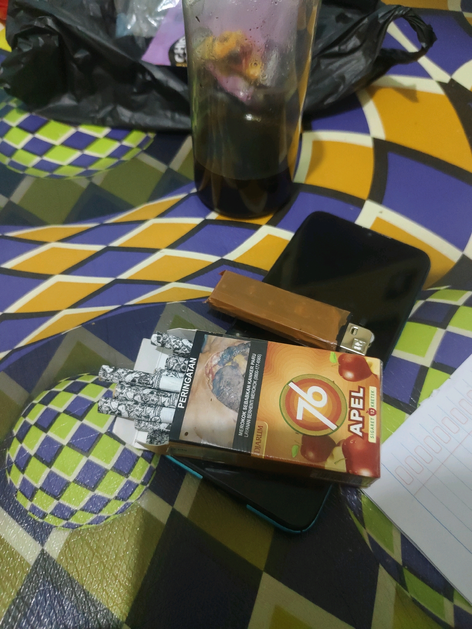 The image depicts a collection of items on a patterned surface, likely a table or bedspread, with a geometric design in shades of blue, yellow, and green. In the foreground, a partially opened pack of "76 APEL" cigarettes is visible, with several cigarettes showing. To the right of the cigarette pack, a lighter with a brown casing lies on top of a black smartphone. Behind the cigarettes, a clear plastic cup filled with a dark liquid is present, next to a crumpled black plastic bag. A sheet of paper with red and blue grid lines and some scribbled text is visible in the lower right corner. The scene is indoors, likely in a residential setting. The image does not provide any specific location details for Sukanagara, Indonesia.