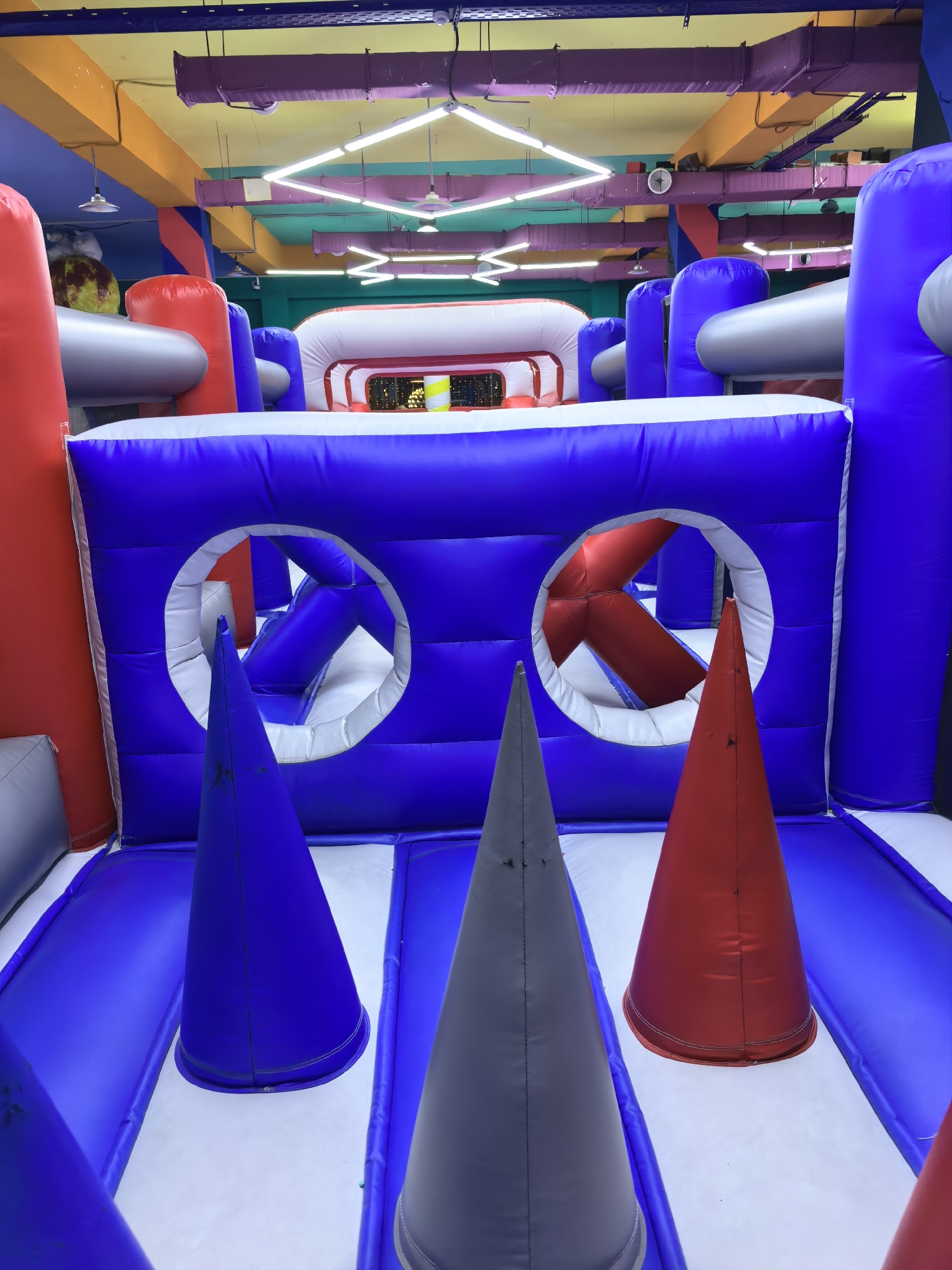 The image depicts an indoor inflatable playground, possibly part of an entertainment center, located in Fergana City, Uzbekistan. The playground features vibrant blue and red inflatable structures, with white accents. Several large, cone-shaped objects in red, blue, and gray are strategically placed on the floor, likely serving as obstacles or targets. Two circular openings are visible in a large blue inflatable wall, allowing passage or views into other sections of the playground. Above, the ceiling shows a colorful industrial design with exposed ventilation ducts painted in yellow and purple, and fluorescent lighting fixtures arranged in a geometric pattern. The overall environment suggests a recreational space designed for children's activities.