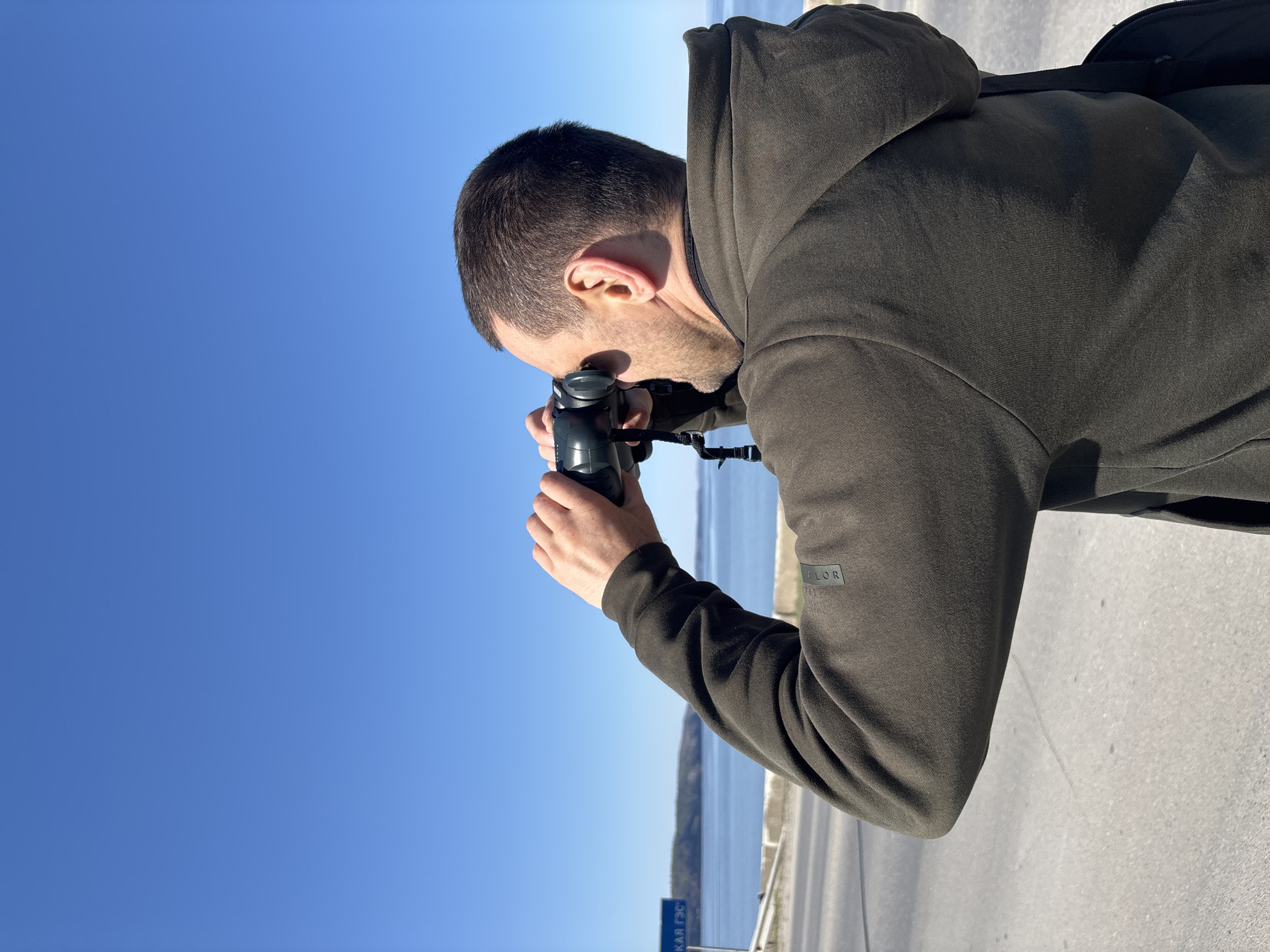 A man stands outdoors, taking a photograph. He wears a dark hooded sweatshirt and a black backpack. The camera in his hands is also black. The background features a bright blue sky, a body of water, and what appears to be a road or sidewalk. A sign with Cyrillic text is visible in the lower left corner, identifying the location as the city of Chaikovsky, Russia. The man is the primary subject, and his action is the focus of the image. The setting is outdoors on a clear day.