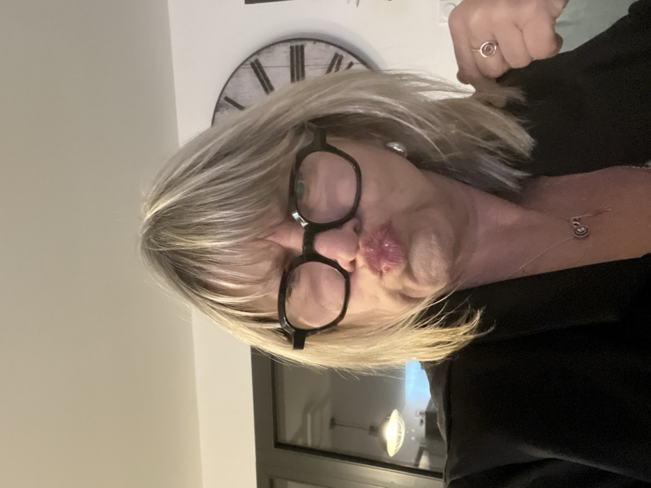 A woman with blonde hair and black-rimmed glasses puckers her lips towards the camera. She wears a black blazer and a delicate silver necklace. A ring is visible on her finger. A wall clock with Roman numerals hangs in the background. A window with a light fixture reflected in it is seen below her face. The overall composition is rotated.