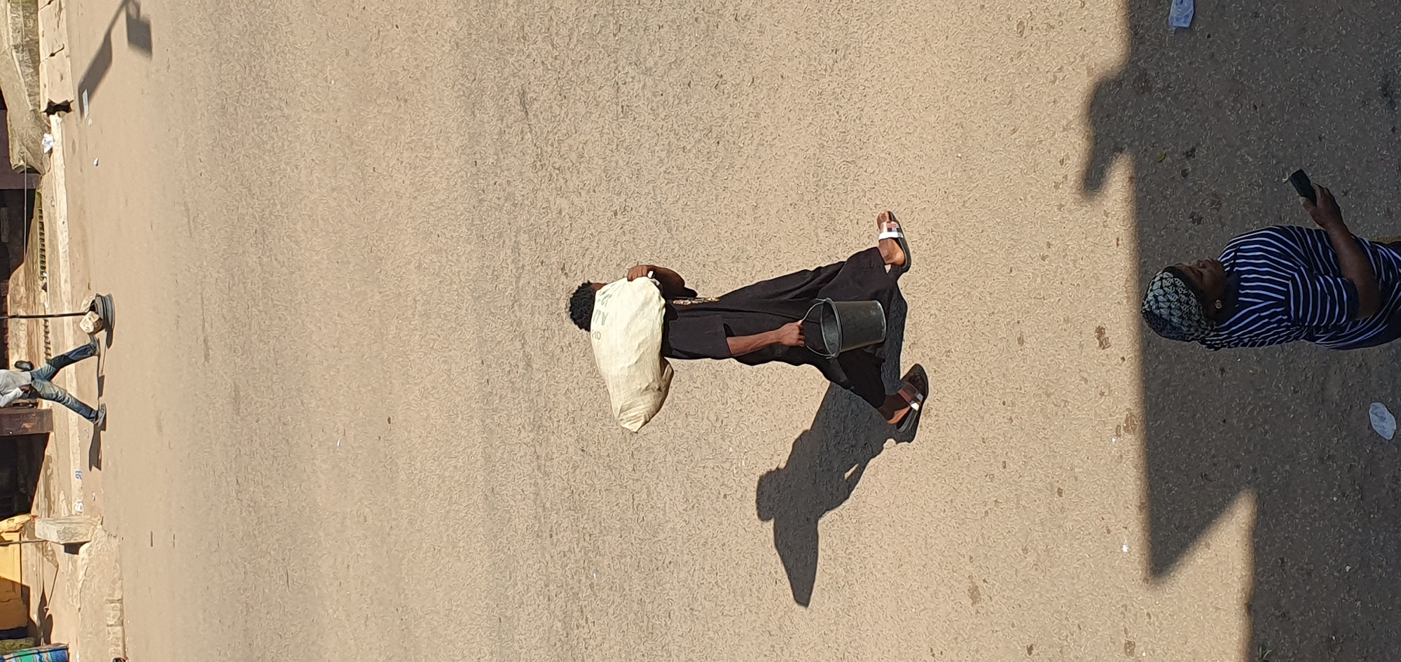 In Bukuru, Nigeria, a person is walking across a paved street, carrying a large, off-white sack balanced on their left shoulder and a dark bucket in their right hand. The person is wearing a black, flowing garment and appears to be wearing sandals. The ground is a textured, light brown asphalt. To the left of the person, a long, sharp shadow is cast on the ground, indicating a strong overhead light source, likely the sun. In the background, a building with a yellow facade and a concrete base is visible. Another person, a woman wearing a striped shirt and a headscarf, is visible in the lower right portion of the image, looking to the right and holding a dark object, possibly a phone. The scene appears to be a typical day in a Nigerian town, with evidence of daily activities and pedestrian movement.
