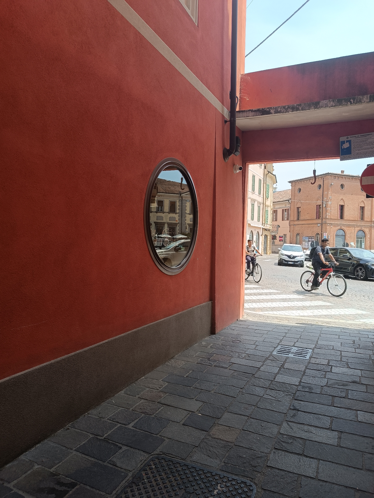 This image captures a sunlit street scene in Casalmaggiore, Italy. The foreground is dominated by a textured red wall on the left, with a dark brown base. A circular mirror on this wall reflects a building with arched windows and a street scene with parked cars and outdoor seating. The right side of the image reveals an archway leading to a paved street. Two men are cycling on the street, with one closer to the viewer on a red bicycle and another further back on a lighter-colored bicycle. Cars are visible on the street, including a white SUV and a black sedan. A crosswalk with white stripes is present on the paved street. The architecture visible in the reflection and beyond the archway suggests a historic European town. The sky is clear and blue. A sign with Italian text and a camera icon is mounted on a pole within the archway.