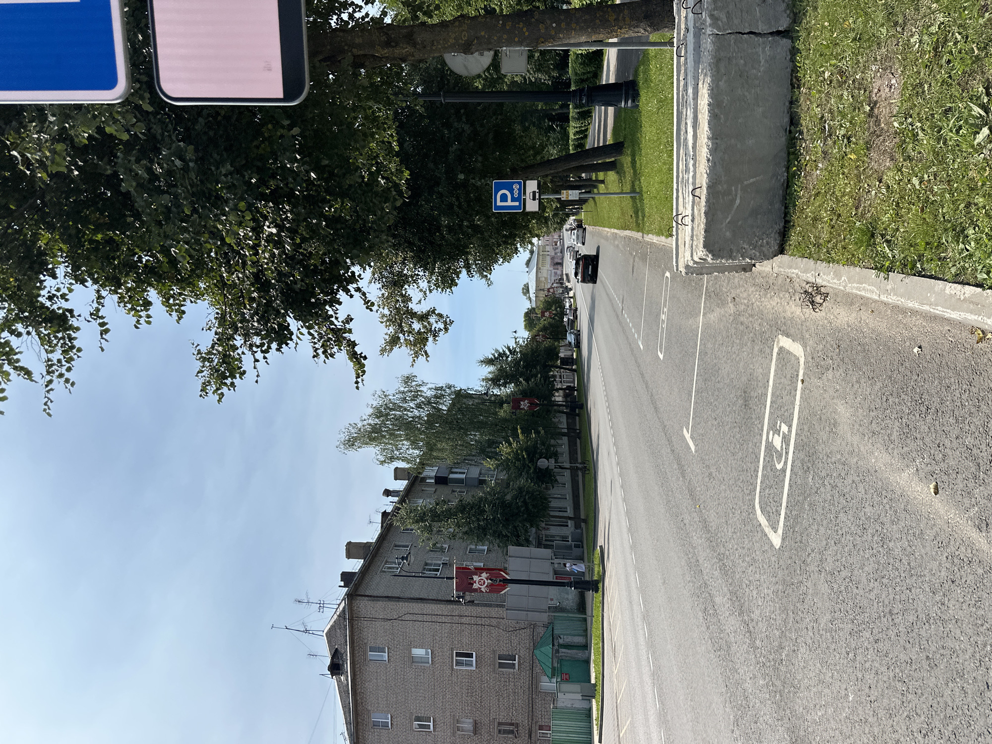 In the city of Selivanovo, Russia, a street scene unfolds under a clear blue sky with scattered clouds. On the left, a multi-story brick apartment building with many windows stands tall. Red banners with a star and hammer and sickle emblem hang from lampposts along the sidewalk in front of the building. The street itself is paved and has white lane markings. On the right side of the street, a row of mature trees with lush green foliage lines the sidewalk. Several concrete blocks are stacked on the grass near the sidewalk. A blue and white parking sign with a silhouette of a car and a time limit is visible on a pole amidst the trees. In the background, other buildings and trees can be seen along the street. Several cars are driving on the road, and a person in white clothing is walking on the sidewalk near the apartment building. The scene appears to be a sunny day in a residential or mixed-use urban area.