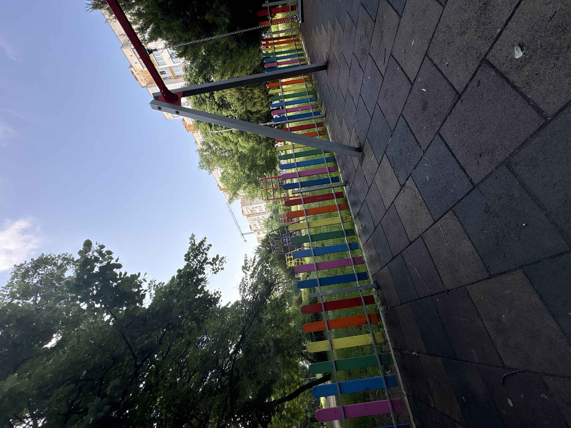 The image captures an outdoor scene, likely a playground or park area, under bright daylight. In the foreground, the ground is covered with dark gray, rectangular paving tiles, possibly rubber safety surfacing, showing some minor wear and a few scattered leaves.
Across the midground, a colorful fence stands out, bordering the paved area. This fence consists of numerous vertical wooden slats, each painted a distinct bright color such as red, orange, yellow, green, blue, purple, and pink, affixed to horizontal metal bars. Behind this vibrant fence, a lush green expanse of grass and abundant trees with dense foliage are visible.
To the upper left, components of a playground structure, specifically robust red and gray metal support beams and a hanging metal chain, are partially visible, suggesting a swing set. In the background, several multi-story apartment buildings can be seen nestled among the trees. A construction crane is also faintly visible in the distance, situated near the buildings.
The sky above is clear and bright blue with only a few wispy white clouds. The strong sunlight and casting of shadows indicate it is daytime, likely mid-morning or afternoon. No people are present in the scene, and there is no visible text. The environment suggests an urban or suburban park setting within Sofia, Bulgaria.
