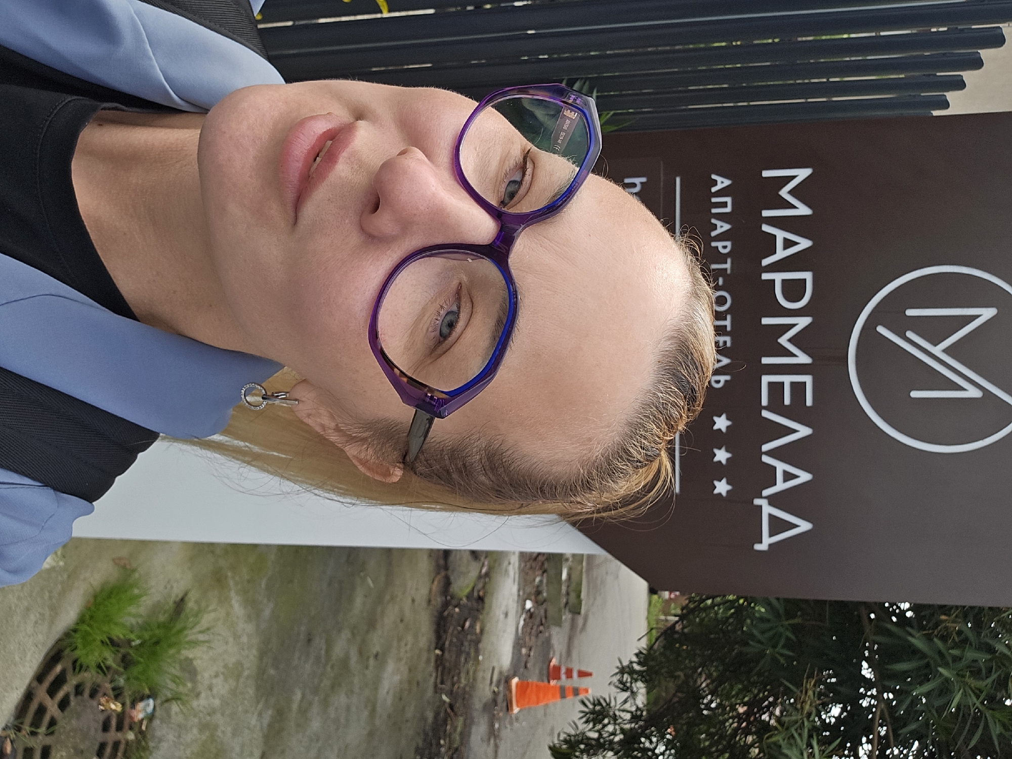 A woman with fair skin and blonde hair is standing in front of a sign in Sochi, Russia. She is wearing purple glasses and a light blue jacket over a black shirt. She has a small, circular earring in her left ear. The sign behind her reads "МАРМЕЛАД апарт-отель," which translates to "Marmalade Apart-Hotel." The sign also features a logo and three stars. In the background, there is foliage, a storm drain, and orange traffic cones.