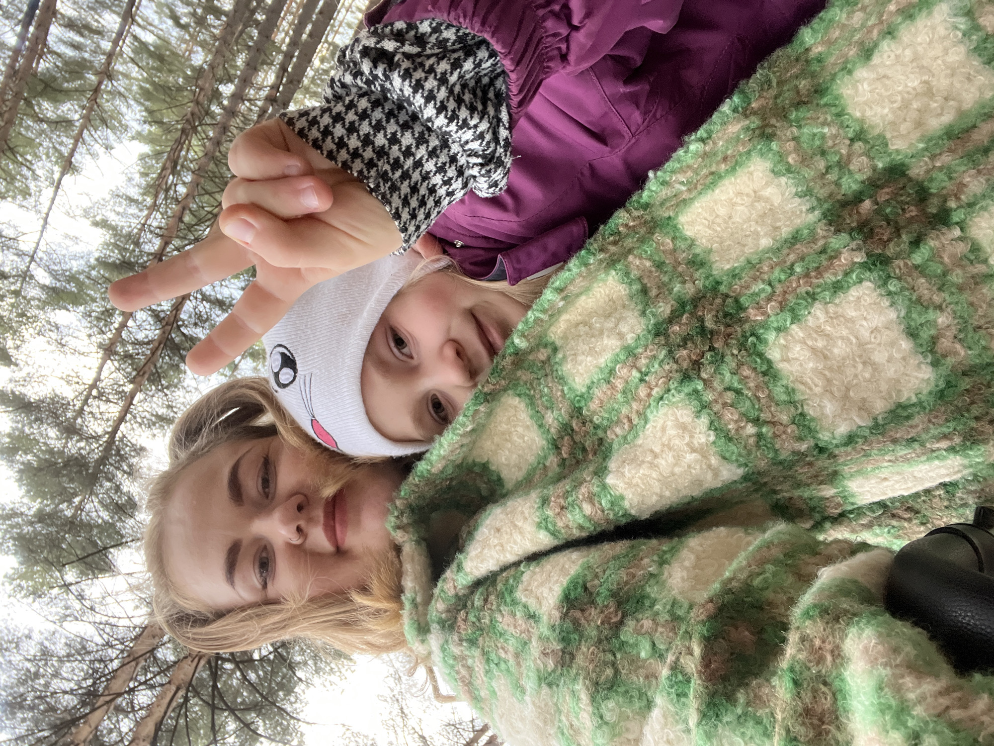 This is a vertically oriented selfie featuring two females outdoors in a pine forest, likely in Chaikovsky, Russia. The image is taken from a low angle, looking upwards towards the tree canopy and a bright sky.
In the foreground on the left, an adult or older teenage female with blonde hair and a nose piercing is visible, looking towards the camera with a subtle smile. She is partially covered by a thick, fuzzy garment with a green, cream, and brown plaid pattern. On her right, slightly behind and above her, is a younger female or child. This child wears a white beanie with cartoon-like ears and facial features, a purple jacket, and a sleeve with a black and white houndstooth pattern. Her right hand is raised, making a peace sign (V-sign) towards the camera.
The background consists of numerous tall, slender pine trees with sparse needles, suggesting a coniferous forest. The sky appears bright and overcast or hazy, indicating it is daytime. The warm clothing worn by both subjects suggests cool weather. The overall mood conveyed is one of casualness and playfulness, with both individuals posing for the camera. There is no visible text in the image.