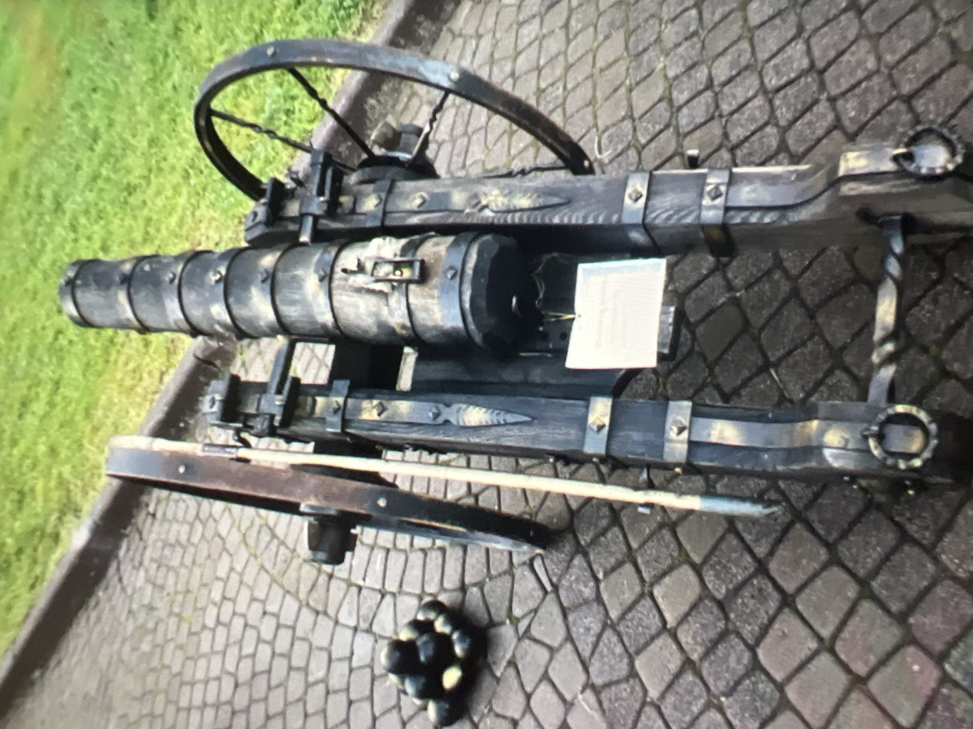 An antique cannon is positioned on a cobblestone path with a grassy area in the background. The cannon is dark in color, with metal bands around its barrel and decorative metalwork on its wooden frame. It has two large spoked wheels. To the left of the cannon, a pile of dark, round cannonballs is visible. A small white sign is attached to the cannon's frame. The scene appears to be outdoors, possibly in a park or historical site. The location in Moscow, Russia, is not explicitly indicated by any visible elements within the image.