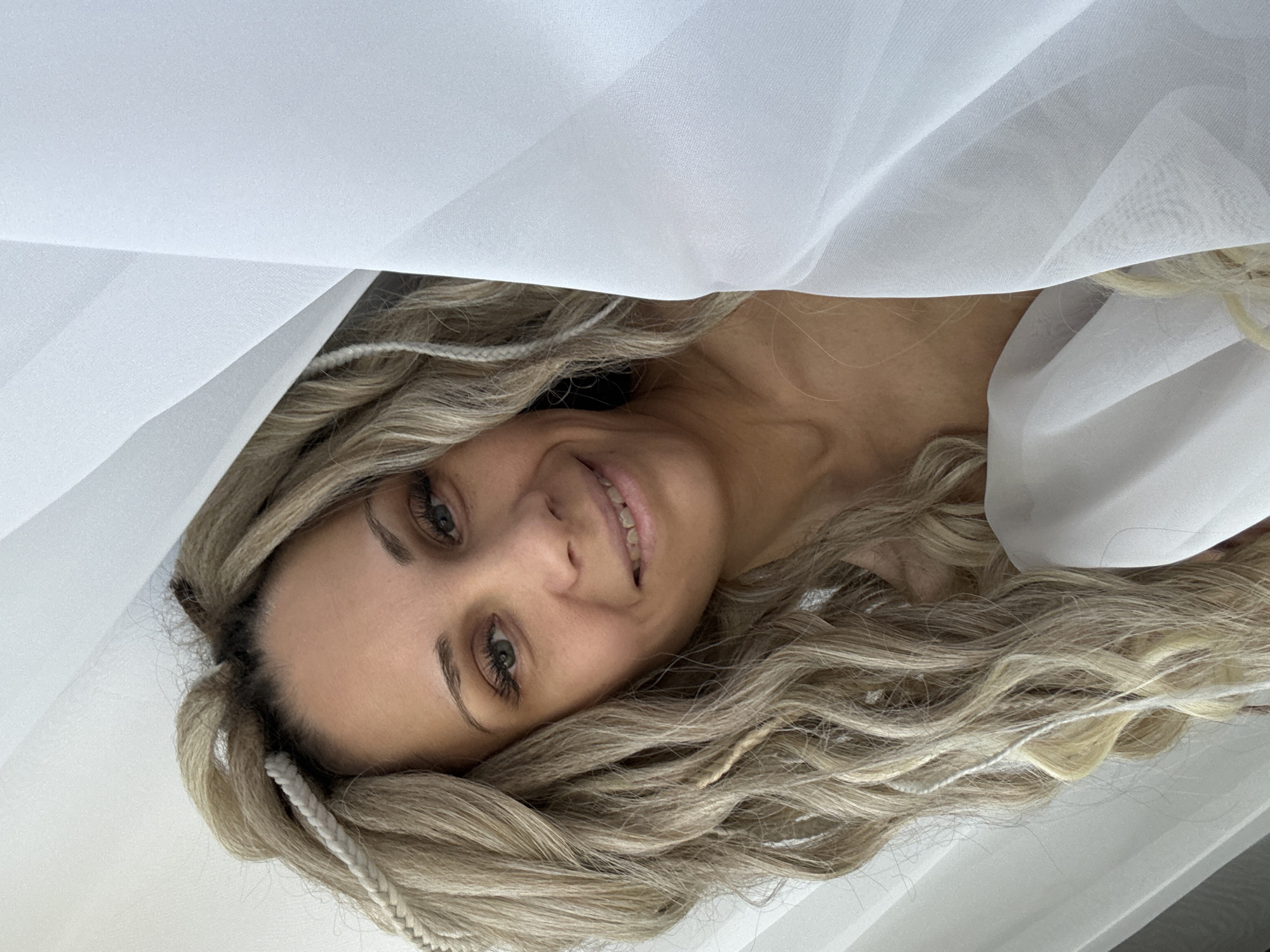 A woman with blonde, braided hair is smiling. She is partially covered by a white, sheer fabric. No information regarding the location is provided.