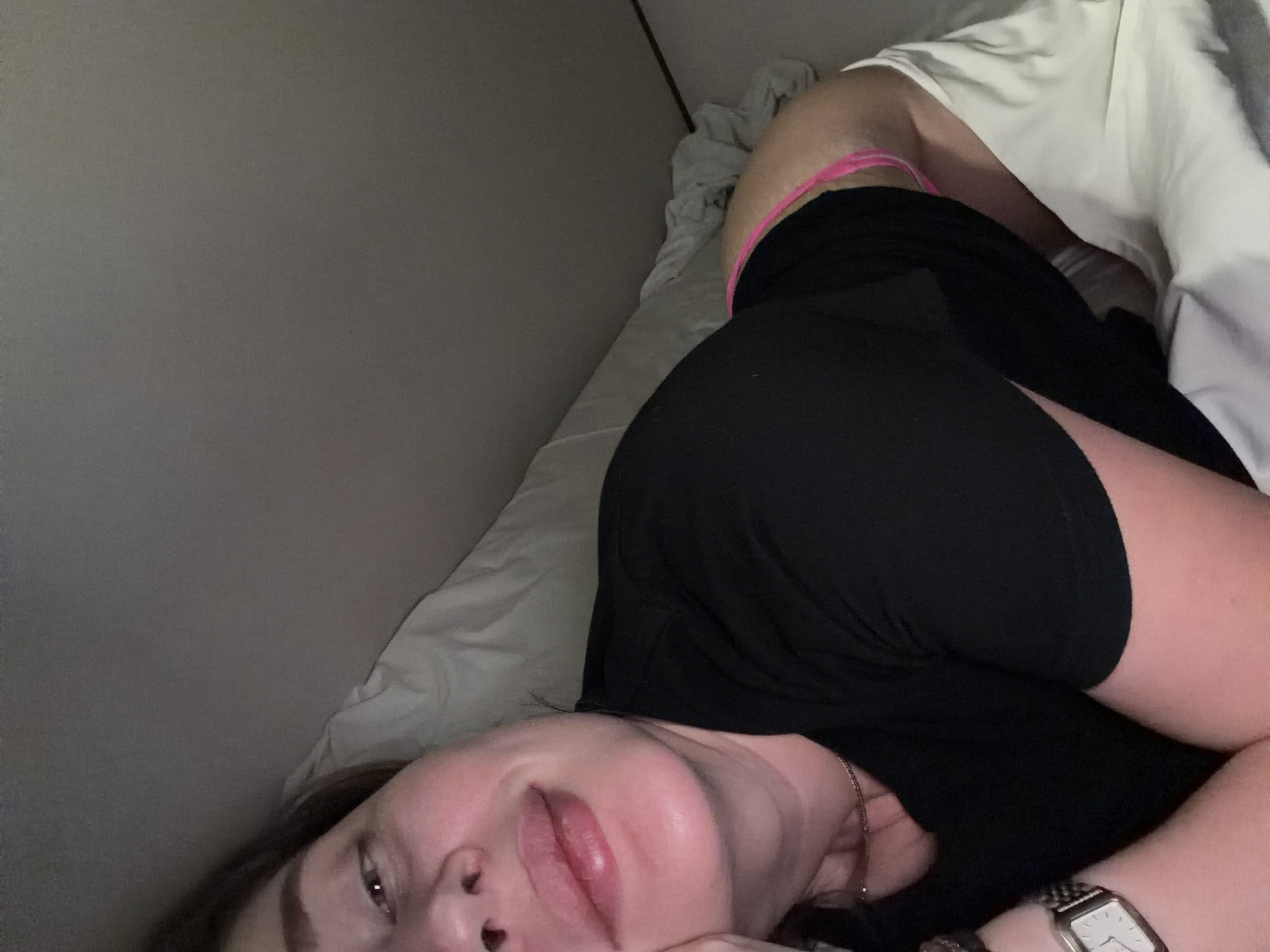 A woman lies on a bed covered with white sheets, partially facing the camera. She is wearing a black shirt and pink underwear. Her head rests on her arm, with a wristwatch visible on her wrist. The background consists of a plain grey wall. The location is not identifiable.