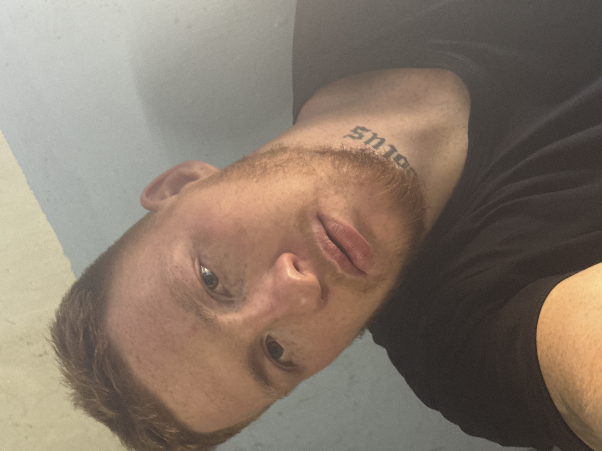 A close-up, head-and-shoulders, selfie-style portrait of a young white male with short, reddish-brown hair and a light beard. He is wearing a black t-shirt and has a tattoo on his neck that appears to read "solus" in gothic lettering. The background is a light blue wall with some textured imperfections. The lighting suggests the photo was taken indoors.