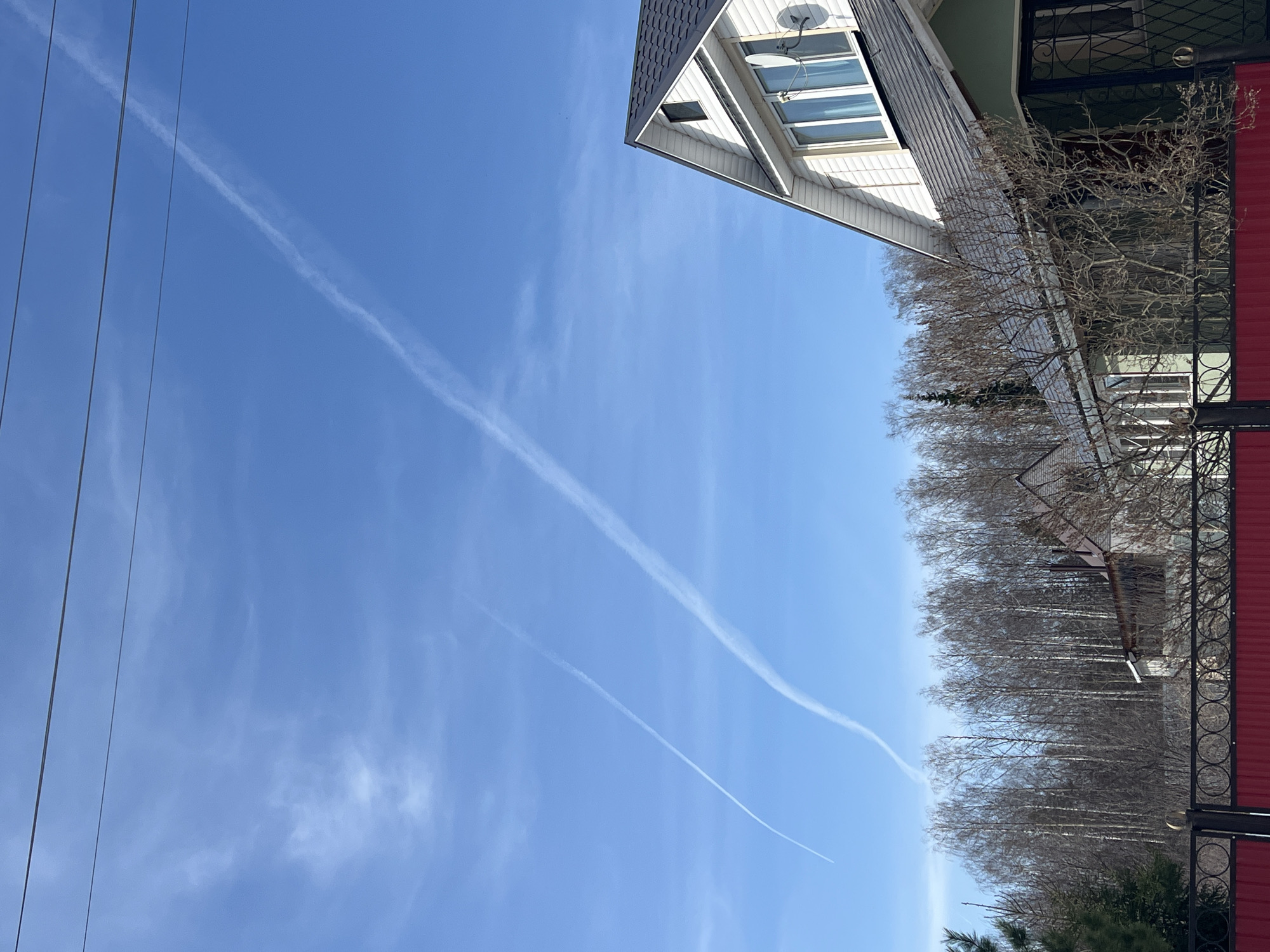 The image shows a daytime scene in Noviy, Russia. The sky is blue with two visible contrails. Power lines are visible in the left portion of the frame. On the right side, there are trees, a house with a satellite dish, and a red metal fence. The angle suggests the photo was taken looking upwards.