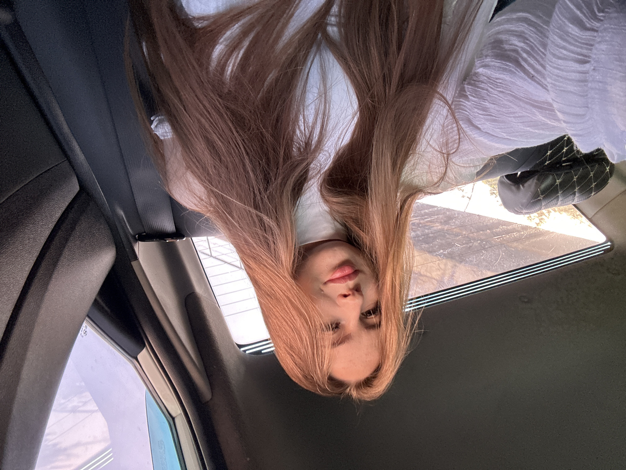 A young Caucasian woman with long, reddish-brown hair and fair skin is pictured from the chest up, inside a vehicle. She is wearing a white turtleneck top and a white ruffled shirt. Her hair is parted in the middle and falls loosely around her shoulders and chest. She is looking directly at the camera with a slight smile. The background shows the interior of a car, including a dark grey headrest with a diamond pattern on the left, a black seatbelt, and a window on the right with a blurred outdoor view. The lighting suggests it is daytime.