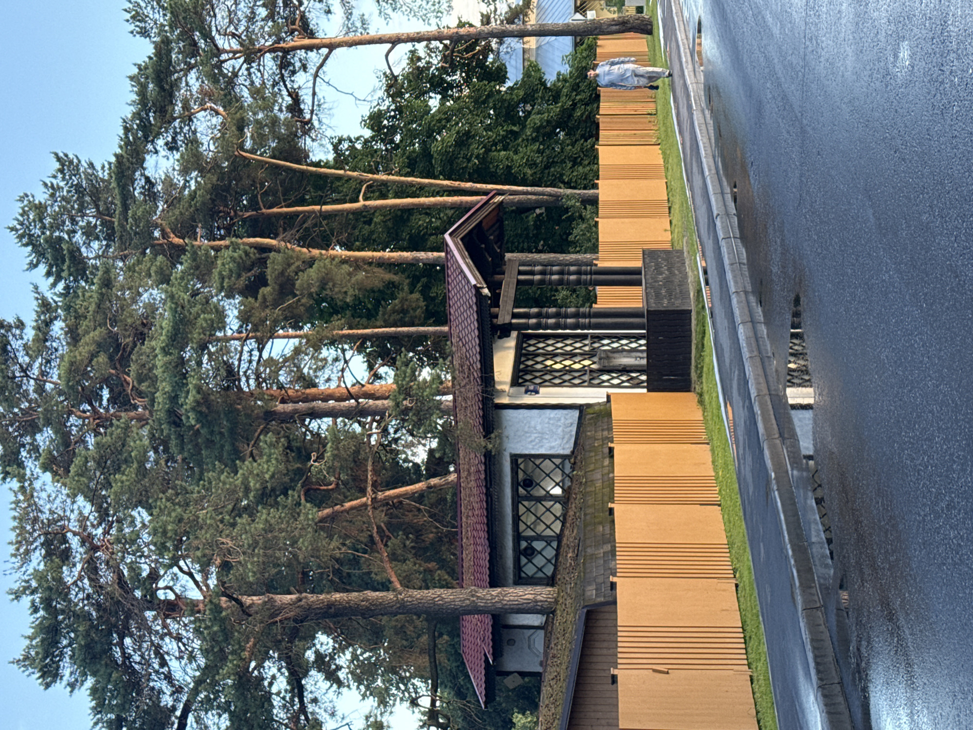 The image depicts a wet asphalt road adjacent to a grassy area and a wooden fence. Behind the fence, a building with a red roof and a patterned window is visible, partially obscured by tall pine trees. The scene appears to be in Moscow, Russia. The time of day is likely late afternoon or early evening, as suggested by the soft light and the wetness of the road, indicating recent rain. A person, dressed in a light blue garment, is standing on the grassy area near the fence, facing away from the camera. The overall atmosphere is calm and somewhat somber due to the overcast weather. There is no visible text in the image.