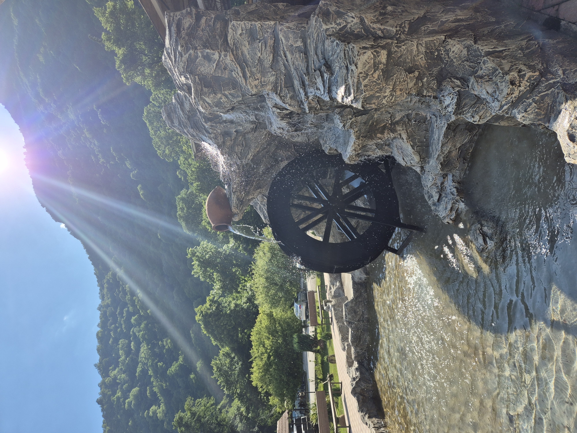 The image depicts a water wheel situated within a rocky, man-made cascade. Water flows from an unseen source above, spilling over the rocks and onto the black spokes of the wheel, creating a dynamic spray of water. The wheel is positioned in a pool of shallow, clear water that reflects the sunlight, causing shimmering patterns on the surface.
Behind the water feature, a lush green mountainside covered in trees slopes upwards towards a bright blue sky. Sunlight streams from the left side of the frame, casting distinct rays and creating lens flare. The overall scene suggests a pleasant, sunny day, likely in the late morning or early afternoon given the angle of the sun.
In the foreground, to the right of the water wheel, a paved walkway is visible, lined with rough-hewn rocks that form the edge of the water channel. Beyond the walkway, there are glimpses of park-like landscaping with benches and trees, and a road with a parked car. A brown, ceramic-like jug is partially visible at the top left, from which water appears to be pouring. The scene evokes a sense of natural beauty enhanced by man-made elements, likely a tourist attraction or a decorative feature in a park or garden. No visible text is present in the image.