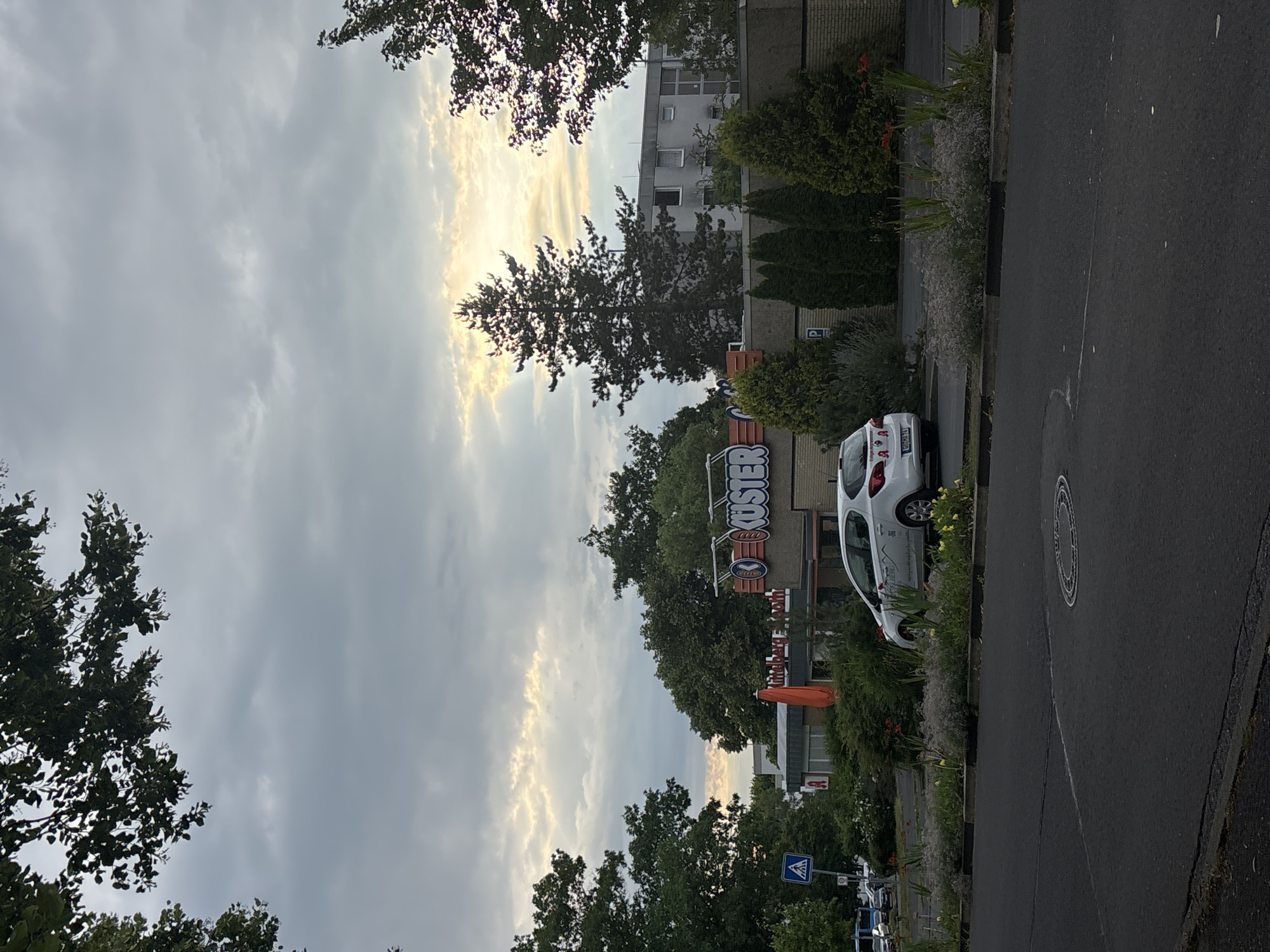 This is a vertical outdoor photograph taken in Göttingen, Germany. The scene depicts a street with a building and vegetation. The sky is overcast with patches of sunlight breaking through the clouds, suggesting it is either early morning or late afternoon.
In the foreground, a paved street is visible, with a curb and a strip of greenery including flowers and bushes separating the street from the building. A white car is parked near the curb. The building in the background appears to be a commercial establishment with a prominent sign. The sign has large, stylized lettering that reads "KUSTER" with additional smaller logos. There are also some orange umbrellas visible near the entrance of the building. Trees with dense foliage are present on both sides of the frame, framing the building and the sky.
A pedestrian crossing sign is visible on the right side of the street. The overall impression of the scene is that of a quiet urban or suburban setting, possibly a commercial district. No people are clearly visible in the frame. The lighting conditions and the overcast sky create a somewhat subdued atmosphere. The time of day is likely during daylight hours, given the brightness of the sky, but the sun's position suggests it's not midday.