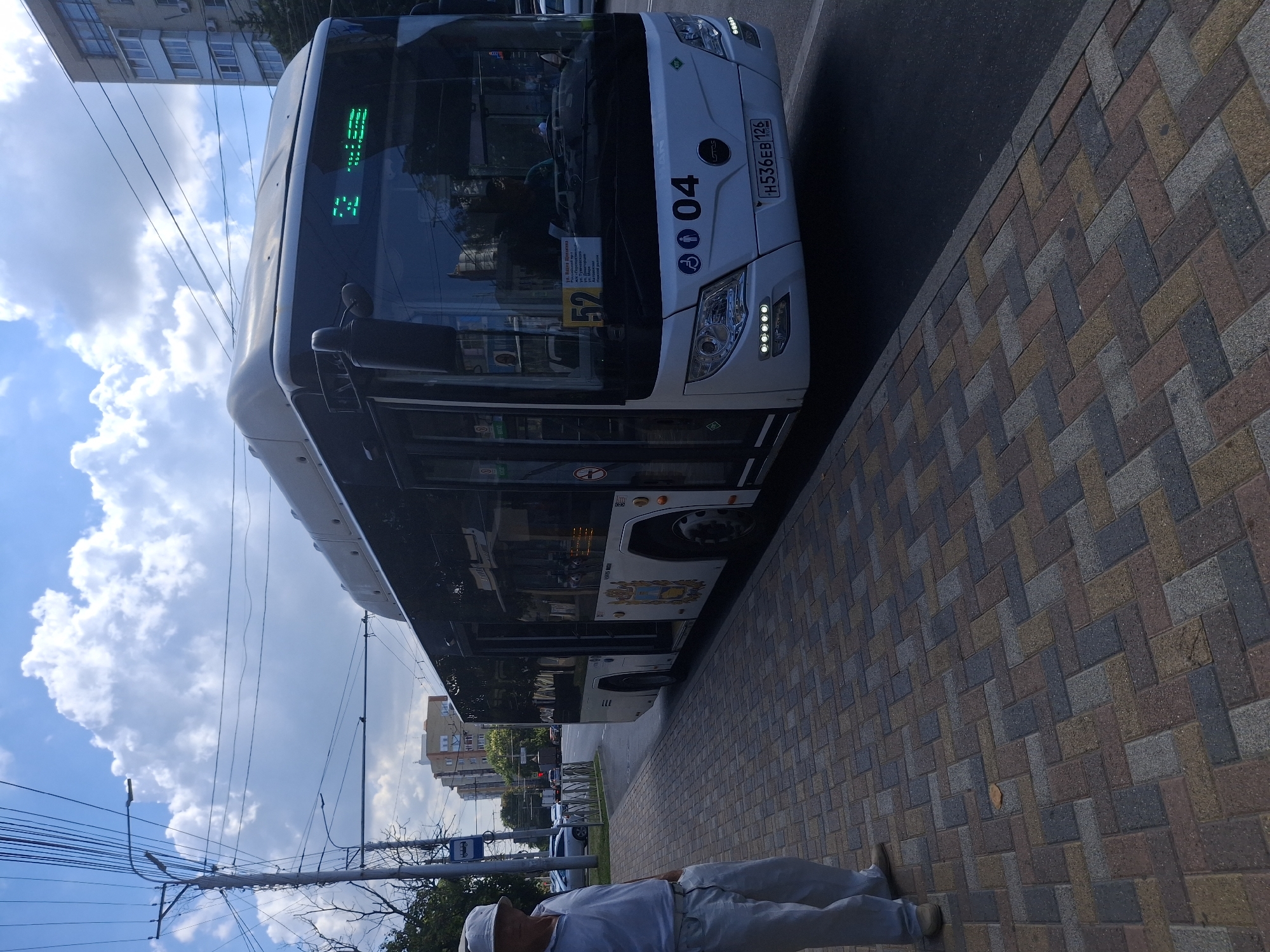 The image shows a white bus with the number "52" displayed on its front. The bus has an electronic sign on the front that reads "52". The bus is parked on a street in Stavropol, Russia. The street is paved with brick pavers. There is a man standing on the sidewalk next to the bus. He is wearing a white hat and a white polo shirt and white pants. The sky is blue with some clouds. There are buildings in the background. A license plate on the bus reads "H536EB 126".