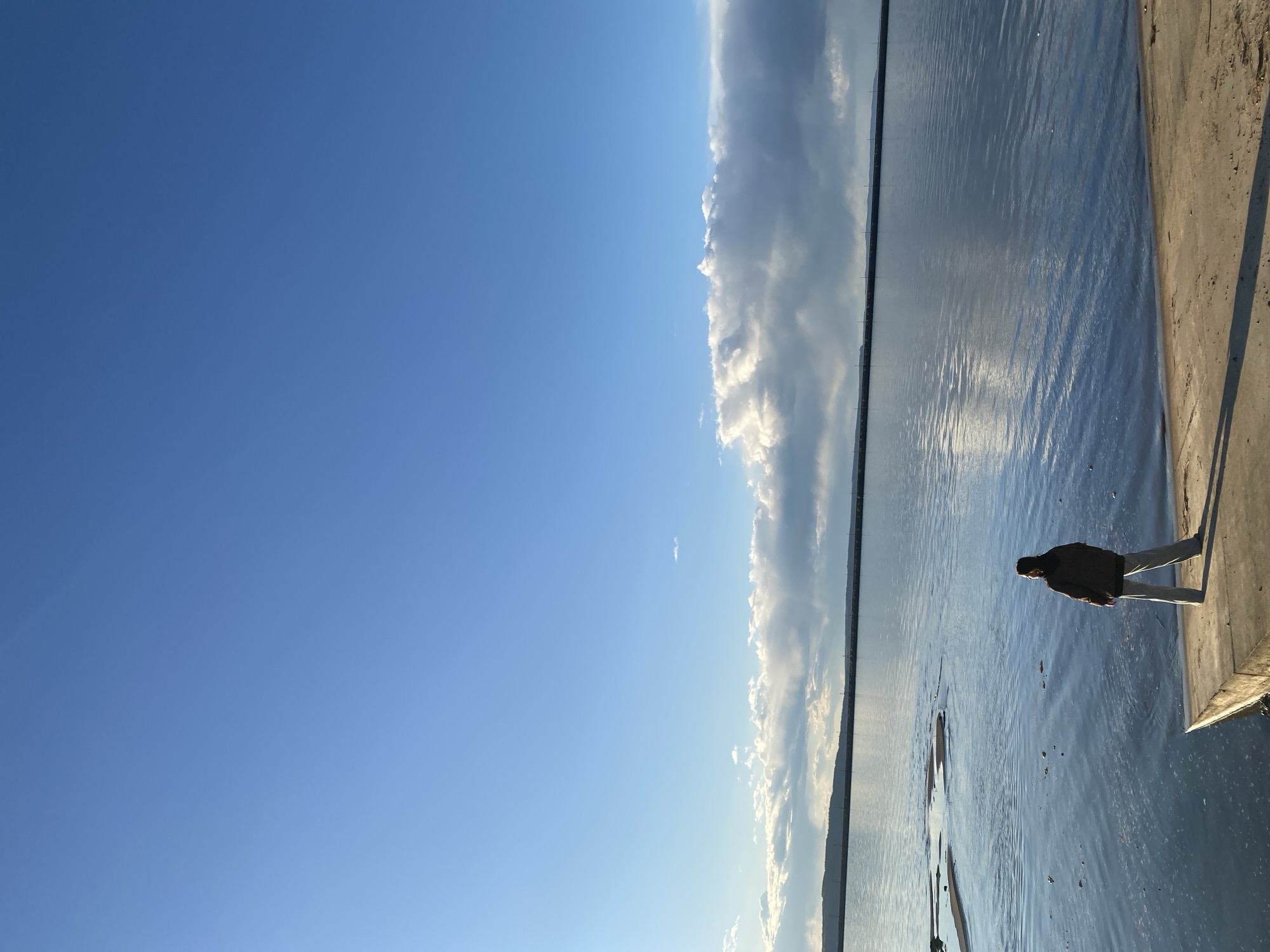 The image shows a scene at the embankment in Chaikovsky, Russia. A person in a dark jacket and light pants stands on the edge of a concrete embankment, looking out over the water. To the left is a clear blue sky, transitioning to a band of clouds reflected in the water. The water appears calm with ripples.