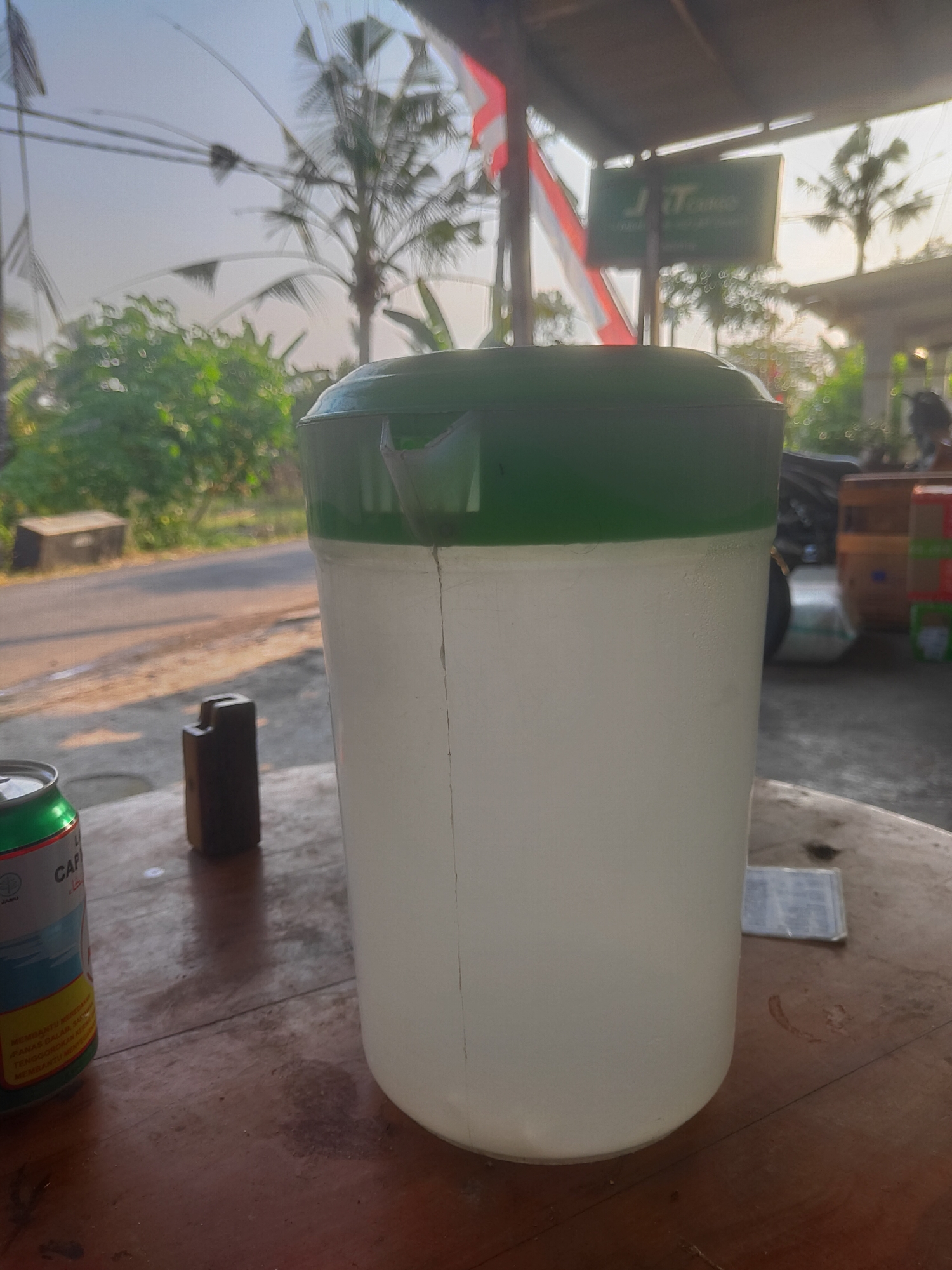 The image depicts a close-up of a large, white plastic container with a green lid, positioned on a wooden surface. The container appears cracked vertically down the front. To the left of the container, a green and silver beverage can is visible. Behind the can and the container, a dark, rectangular object stands upright. The background suggests an outdoor setting, possibly a roadside stall in Serut, Indonesia, with palm trees, greenery, and a road visible. A sign with the text "JiTono" is also present in the background, along with what appears to be a motorcycle and stacks of goods. The lighting indicates it is either early morning or late afternoon.
