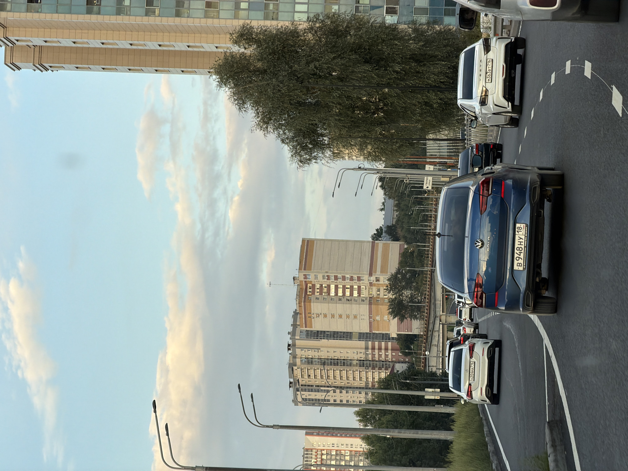 The image shows a street scene in an urban environment. A multi-lane road curves to the right, with several cars driving in the same direction. The cars include a white Audi, a dark blue Volkswagen, and a white Lada Vesta. In the background, there are tall, modern buildings, some with many windows, and several streetlights. Lush green trees line the side of the road. The sky is a pale blue with scattered white clouds, suggesting it is daytime. The road markings are visible, including white dashed lines and solid white lines. There are traffic signs, including one with Russian text. The overall impression is of a busy city street during daylight hours.