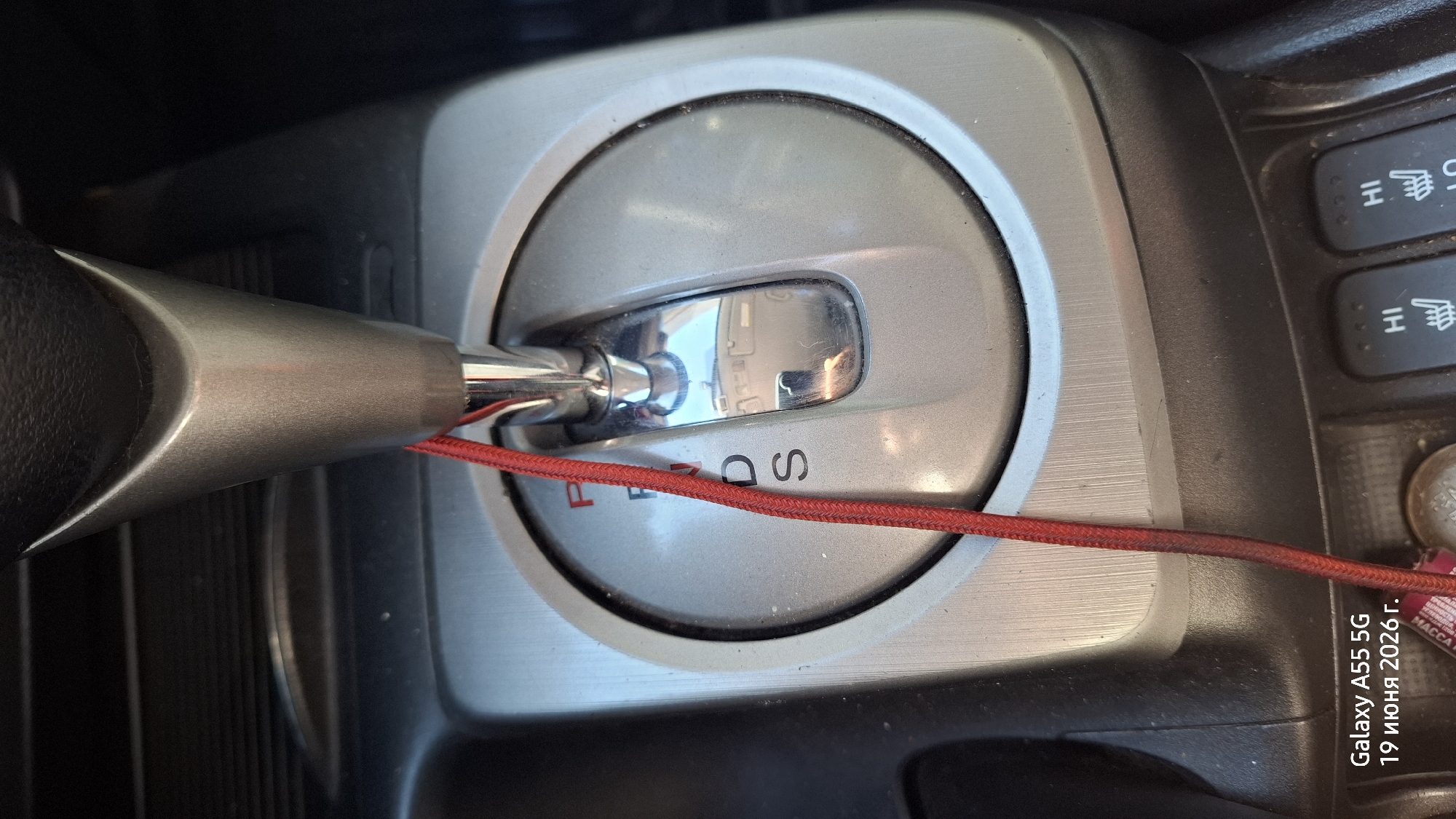 This is a close-up, top-down photograph of a car's automatic gear shifter and center console. The gear shifter, with a silver and black handle, is positioned in the "P" (Park) setting, indicated by a red line that also highlights the "D" and "S" (Drive and Sport) options. The gear selector is surrounded by a brushed silver trim with a circular design. To the right of the gear selector, a row of dark gray buttons with illuminated symbols (heating elements and "HI") are visible, suggesting the presence of heated seats. A small coin and a portion of a red cable are also present on the console. The overall setting appears to be the interior of a vehicle, likely parked, with the lighting suggesting it could be daytime. Text overlaid on the image, likely from a phone's timestamp, reads "Galaxy A55 5G 19 июня 2026 г." and "MACCA". The context provided indicates the location is Rostov-on-Don, Russia. There are no people visible in the image.