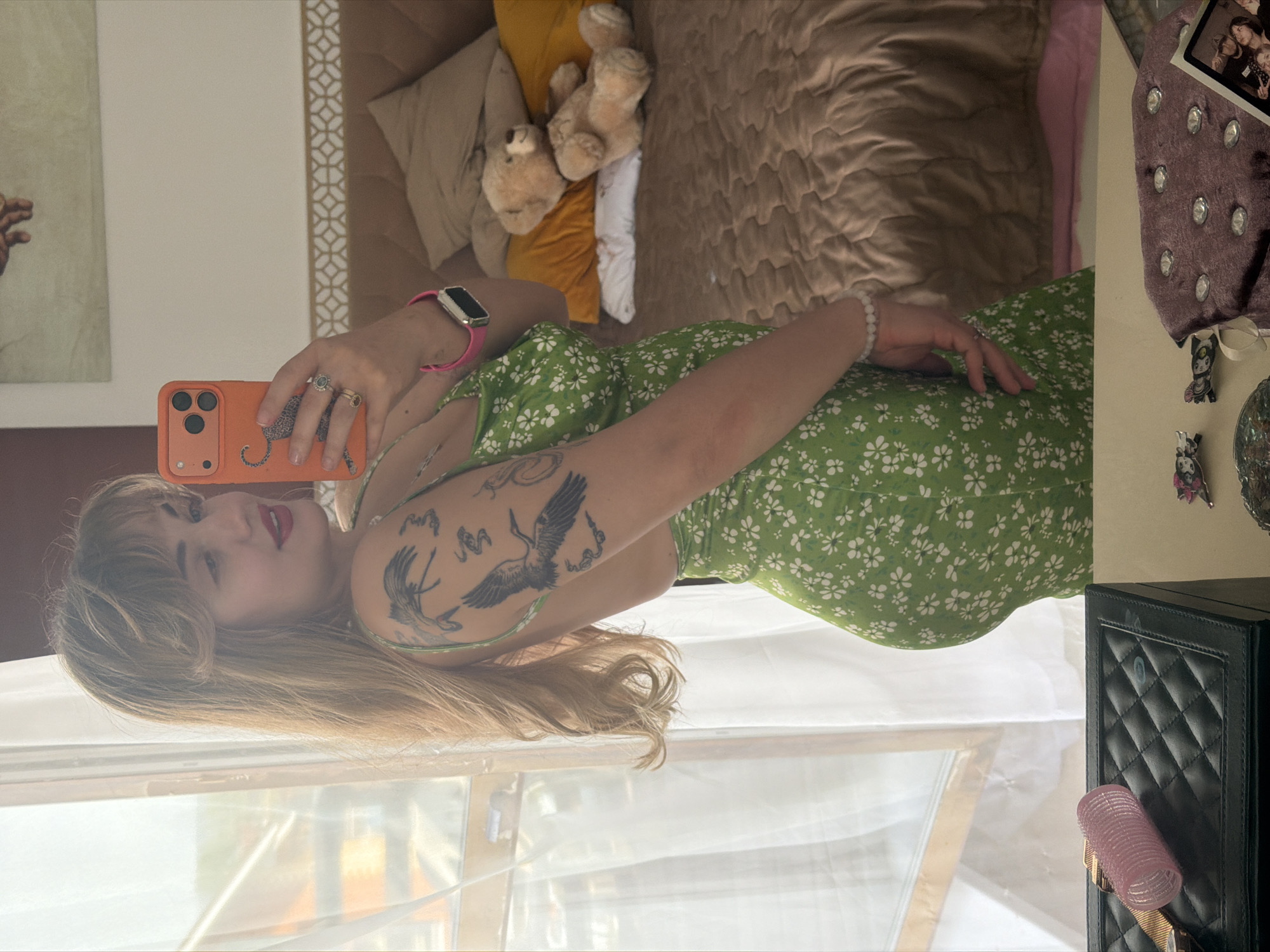 The image shows a young woman taking a selfie indoors, likely in a bedroom setting. She is wearing a green floral dress and has tattoos on her arm. She is holding an orange phone with a case. The background includes a bed with multiple pillows and blankets, a picture frame, a vanity table with items, and a window with translucent curtains.
I cannot pinpoint the specific location in Большая Шильна based on this image alone. It lacks any distinctive landmark or identifying geographical information.