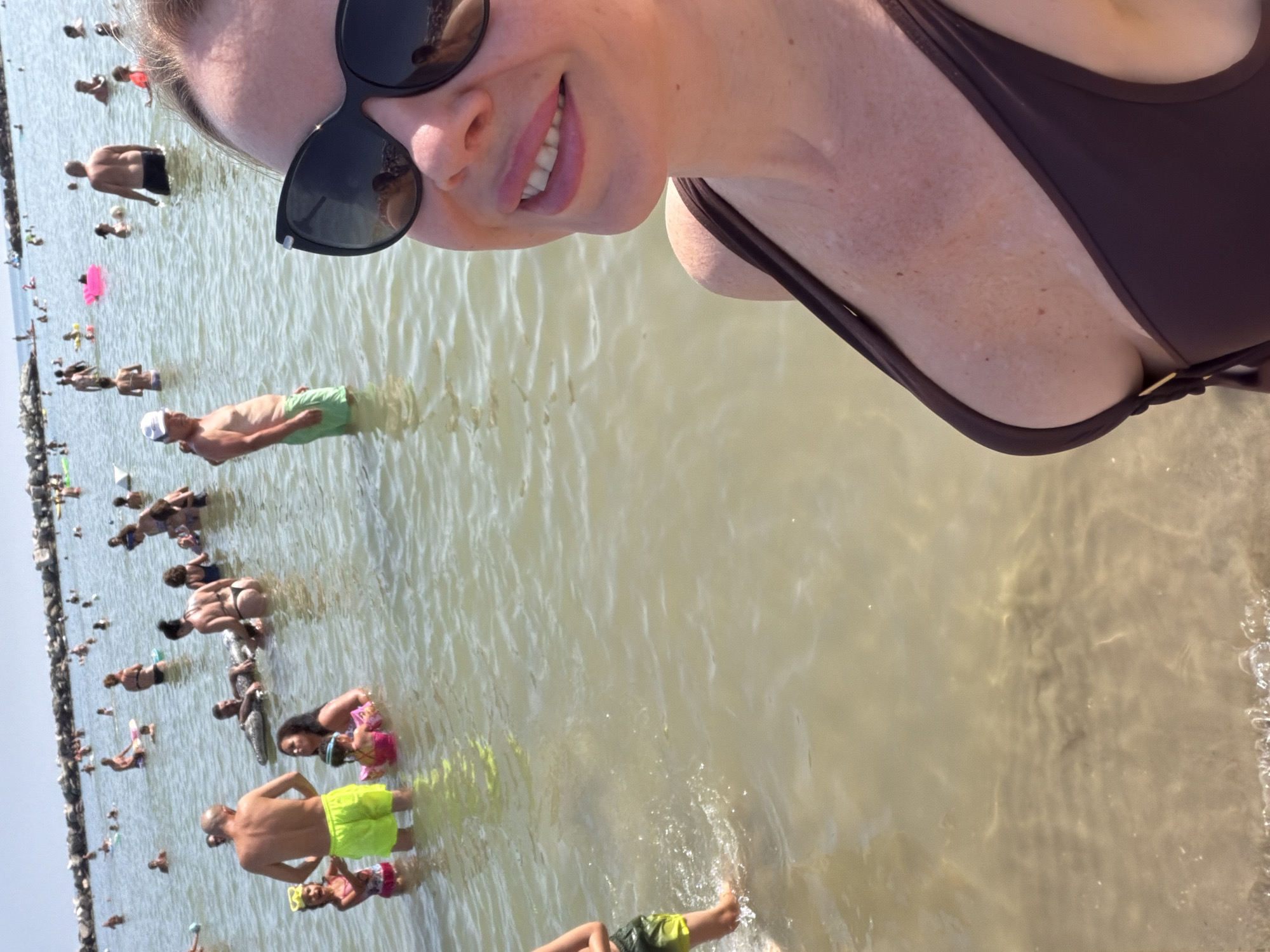 In Gatteo, Italy, a woman in a brown bikini takes a selfie at the beach. She is smiling and wearing large black sunglasses. Behind her, many people are in the shallow, murky water. Some are swimming, while others are standing or sitting in the water. Children are visible playing and wearing swimsuits. The background shows a rocky breakwater and a clear sky.