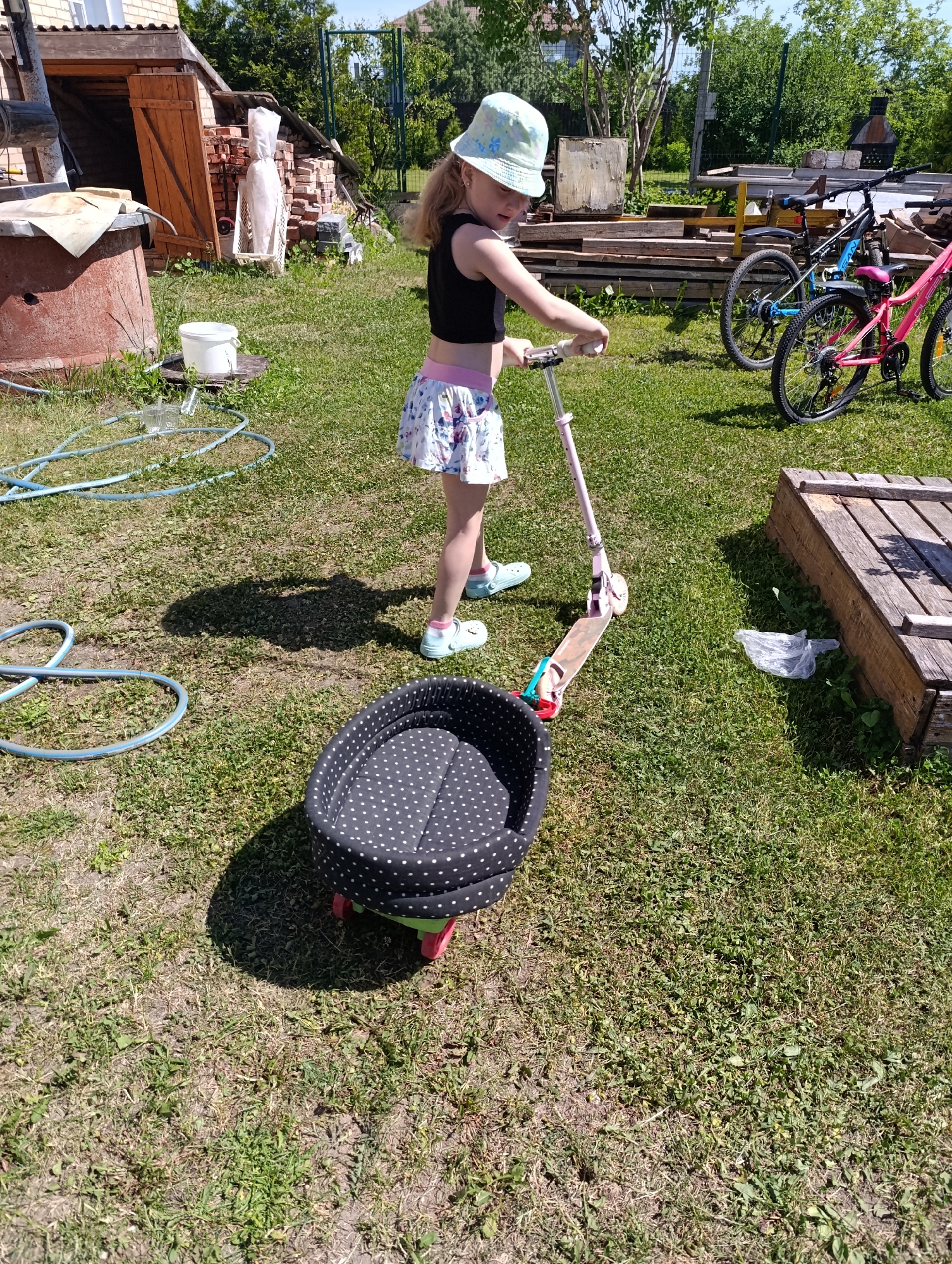 A bright, sunny outdoor scene depicts a residential or semi-rural backyard in the city of Unknown, Unknown, with a young child and various scattered objects on a grassy, uneven terrain.
A fair-skinned female child, approximately 5-8 years old, stands centrally, holding the handlebars of a pink scooter. She wears a light blue bucket hat with a subtle floral pattern, a black sleeveless crop top, a white floral-patterned skirt with a pink waistband, and light blue closed-toe clogs worn with pink socks. Her gaze is directed downward towards the ground or the scooter's path.
Immediately in front of the child and the scooter is an oval-shaped pet bed, covered in black fabric with white polka dots and mounted on a lime green base with small pink wheels. The scooter's front wheel is positioned near the pet bed, suggesting an imminent interaction or maneuver.
To the left of the scene, a blue garden hose is loosely coiled on the grass. Further left, a white plastic bucket rests on a small wooden plank or pallet, with a clear glass jar nearby on the grass. To the right of the child, a small crumpled transparent plastic bag lies on the grass beside a rectangular wooden structure, possibly a raised garden bed frame or a large crate, lying horizontally.
In the background on the left, a large, rust-colored metal container or well cover is partially visible. Behind it, a small wooden shed with a partially open wooden door stands, flanked by a stack of red bricks against which a white, wrapped object leans. A light-colored brick building is visible behind the shed, and dense green foliage frames the scene. On the right side of the background, two bicycles, one blue and one pink mountain bike style, are parked upright. Behind them, a substantial pile of various wooden planks and lumber is stacked. Further back, a metal structure, possibly a grill or outdoor stove, is visible near trees. A green wire fence marks the property boundary in the far distance.
The ground consists of green grass interspersed with patches of dry grass and bare earth. Strong sunlight casts clear shadows, notably the child's elongated shadow and the distinct shadow of the pet bed.