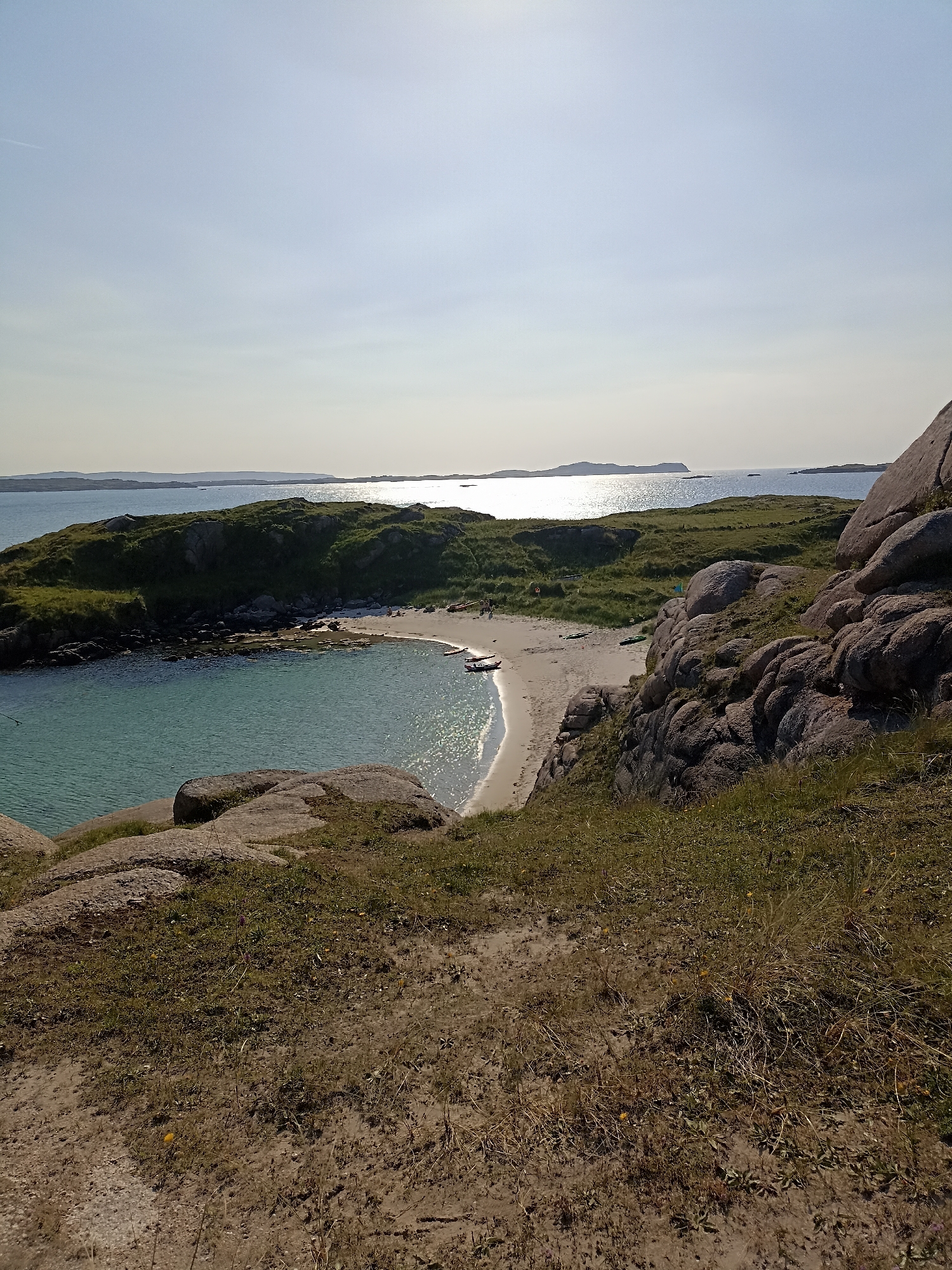 This is a photograph of a small, secluded cove on the coast of Carnboy, Ireland, under a clear sky. The foreground features dry, sandy ground with sparse, brown vegetation and large, textured grey boulders. The middle ground reveals a crescent-shaped sandy beach meeting calm, turquoise water. Several small boats, including what appear to be kayaks, are beached on the sand. A few individuals are visible on the beach. The background consists of a low, green, grassy island with rocky outcrops, and beyond that, the expansive, shimmering sea under a bright sun, with distant islands silhouetted against the horizon. The overall impression is of a peaceful, natural coastal landscape.