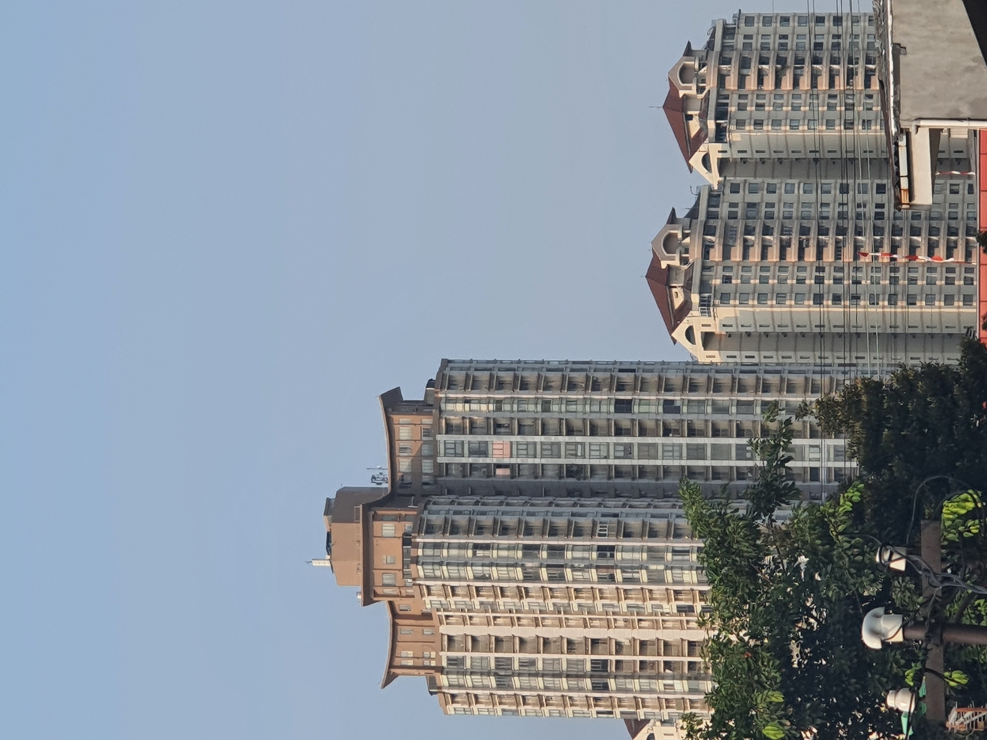A telephoto shot captures a cluster of modern, high-rise apartment buildings against a clear, pale blue sky in West Jakarta, Indonesia. The architecture is characterized by tiered roofs, balconies with white railings, and numerous windows. The tallest building on the left features a distinctive curved roofline and a beige, cylindrical structure at its peak. To its right, two symmetrical buildings with prominent red-tiled roofs and arched detailing are visible. In the foreground, lush green trees obscure the lower portions of the buildings, and power lines with ceramic insulators are present. The scene appears to be a residential area bathed in the warm light of a sunny day.