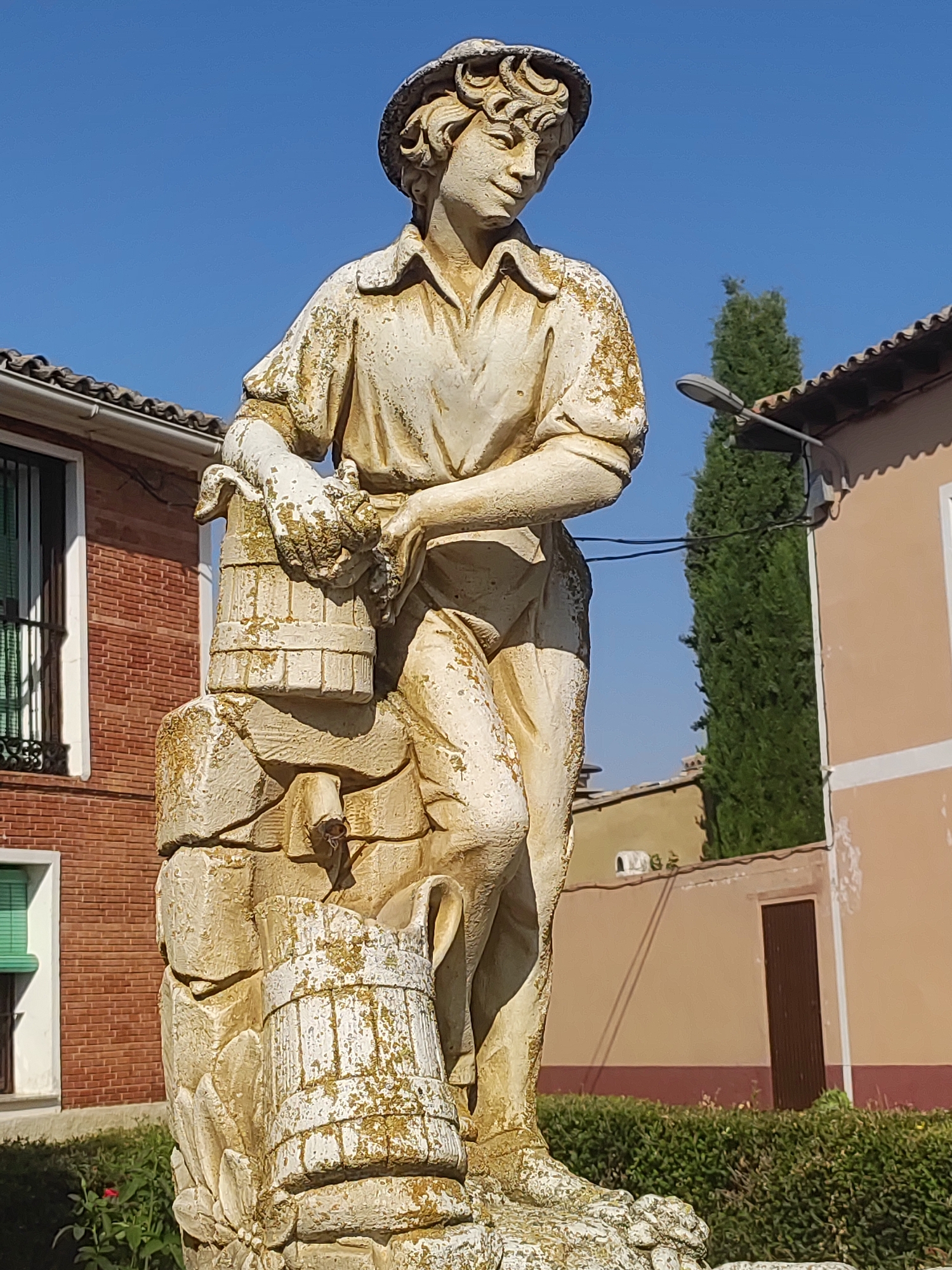 The image shows a weathered stone statue of a young man wearing a hat and a collared shirt, holding a bucket. The statue is made of light-colored stone and shows signs of age and weathering, with patches of green moss on its surface. The man has his right arm bent, with his hand resting on the top rim of the bucket, and his left arm is bent as well, holding the bucket by its handle. The bucket appears to be made of wood or stone and is cylindrical in shape. The statue is positioned in an outdoor setting, with buildings and vegetation in the background. To the left of the statue, a red brick building with a window is visible. To the right, a light-colored building with a dark-colored door and a tall, slender evergreen tree are present. The sky is clear and blue. The statue is likely a fountain or a decorative piece in a public space, potentially a plaza or garden. The location is specified as Autillo de Campos, Spain.