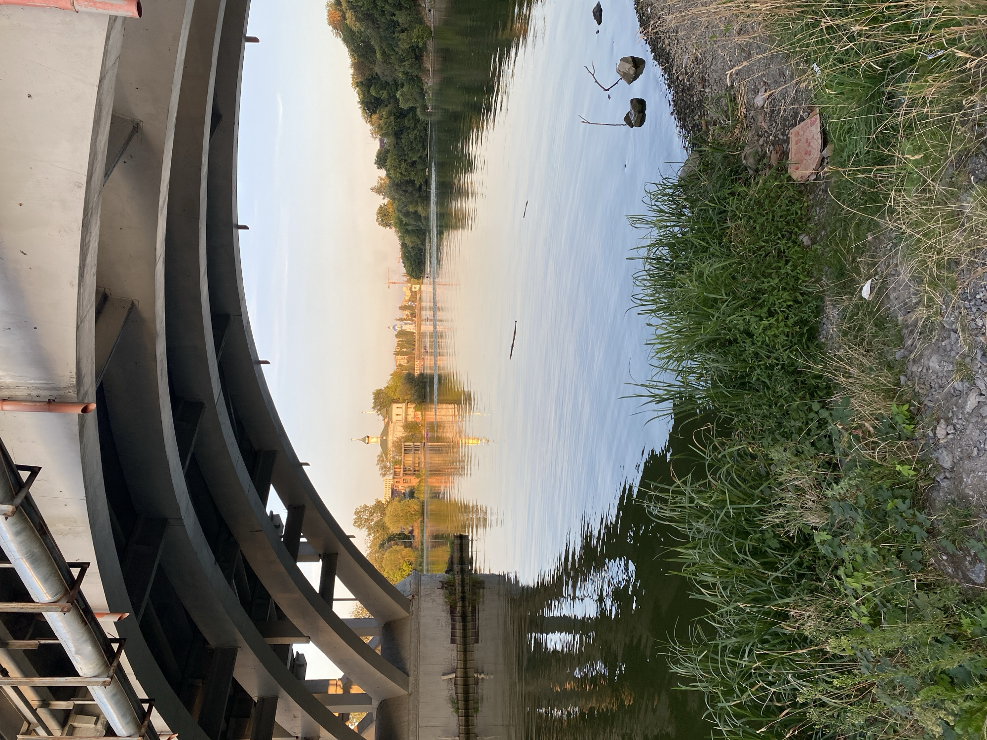 The image captures a view from under a bridge in Vinnytsia, Ukraine, looking out over a wide river during the late afternoon or early evening. The bridge's underside, with its grey concrete and metal structures, dominates the foreground. The river's surface is calm, reflecting the sky and the tree-lined opposite bank, as well as buildings in the distance. On the far bank, several structures are visible, including what appears to be a church with a golden dome, and other buildings nestled amongst dense green trees. The sky is a soft gradient of pale blue and warm yellow, suggesting sunset. The riverbank in the immediate foreground is overgrown with tall green grass and weeds, with some rocks and debris visible. There are a few people faintly visible on the river in small boats or kayaks, and some on the distant bank, indicating recreational activity. The overall atmosphere is peaceful and scenic, with the urban landscape subtly present in the background.