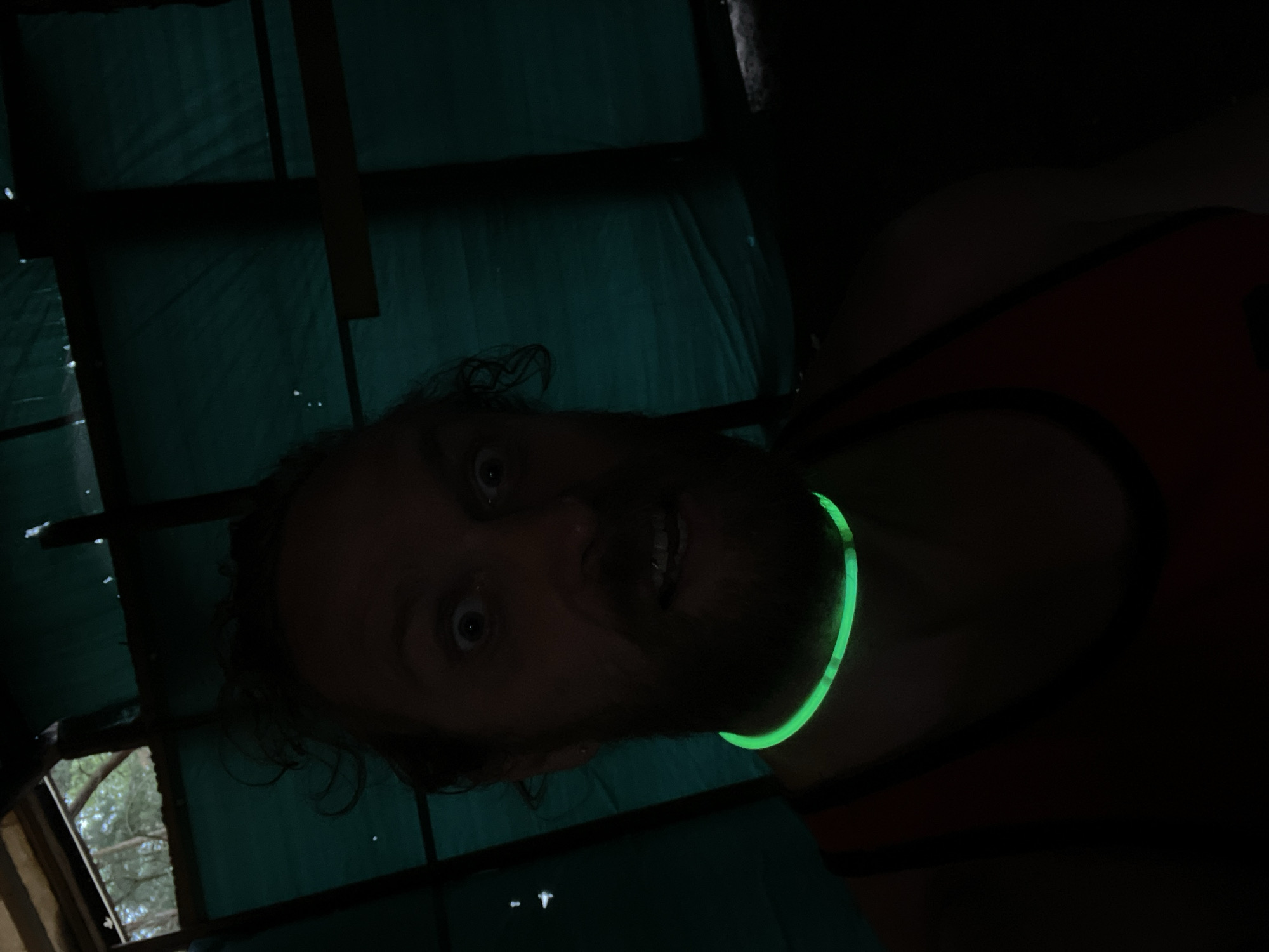 The image is a close-up, self-taken photograph of a man inside a dark structure. The man has a beard and wild, curly hair. His eyes are wide open, and his mouth is slightly ajar, revealing his teeth. He is wearing a neon green glow necklace that is brightly illuminated. The background appears to be some sort of makeshift shelter, with a greenish tarp-like material covering the walls and ceiling, and wooden beams supporting the structure. Glimpses of trees and the sky are visible through an opening at the top left of the frame. The overall lighting is dim, with the glow necklace providing the primary light source for the foreground. The location is described as being in the city of Luzhki, Russia.