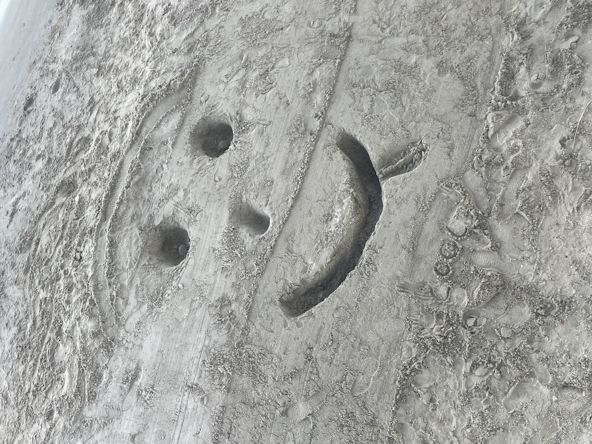 A close-up shot of light gray sand shows a smiley face drawn into the surface. The eyes are two small, circular depressions, while the mouth is a curved indentation. The sand around the drawing is textured with footprints and other disturbances. The image was probably taken at a beach near Menderes, Turkey.