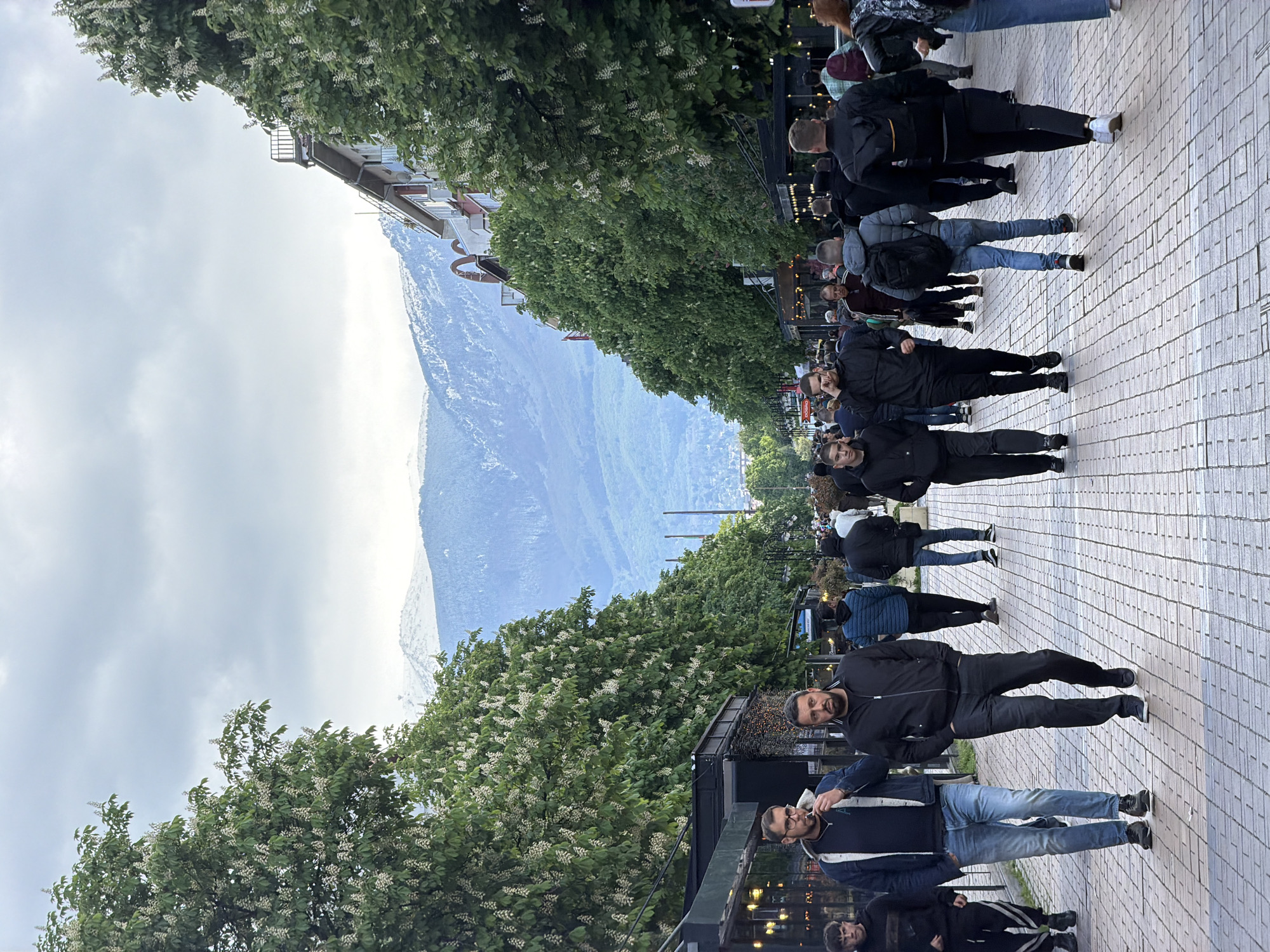 The image captures a bustling street scene in Sofia, Bulgaria, framed by lush trees blossoming with white flowers. A large, snow-capped mountain looms in the background. A crowd of people, mostly dressed in dark clothing, stroll along a paved walkway. Two men are standing near the front of the crowd. The man closer to the camera is wearing a black jacket and jeans, while his companion is dressed in a black jacket. The atmosphere suggests a popular location within the city, likely a pedestrian zone with scenic views.