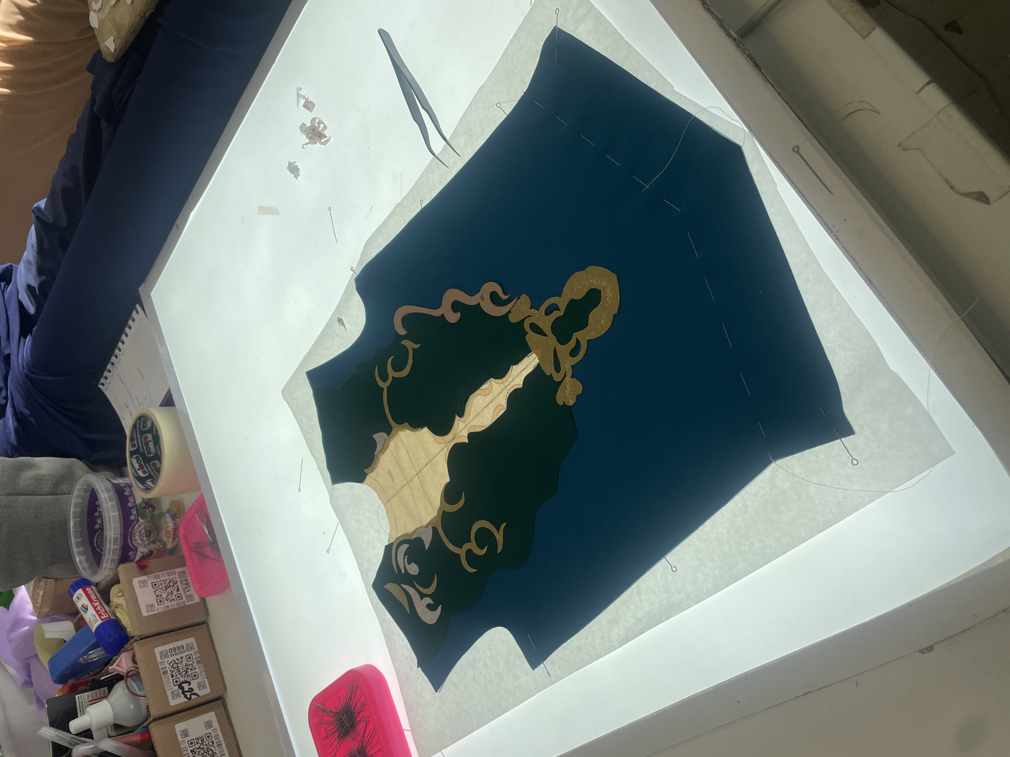 An overhead shot captures a crafting project on a light table. A dark blue garment, possibly a vest, lies flat on the table, covered by a layer of tissue paper and a pattern. Gold and dark green applique elements are partially attached to the garment, creating an ornate design on the front. Sewing pins hold the fabric and pattern in place. To the left, a pink pincushion is filled with pins, and several small cardboard boxes with QR codes are stacked nearby. A glue stick, a tube of white liquid, and a roll of tape are also visible on the edge of the table. In the background, a person is seated, with their legs extending into the frame. The scene appears to be an indoor crafting space, likely a home or workshop, with no specific city or geographical markers present.