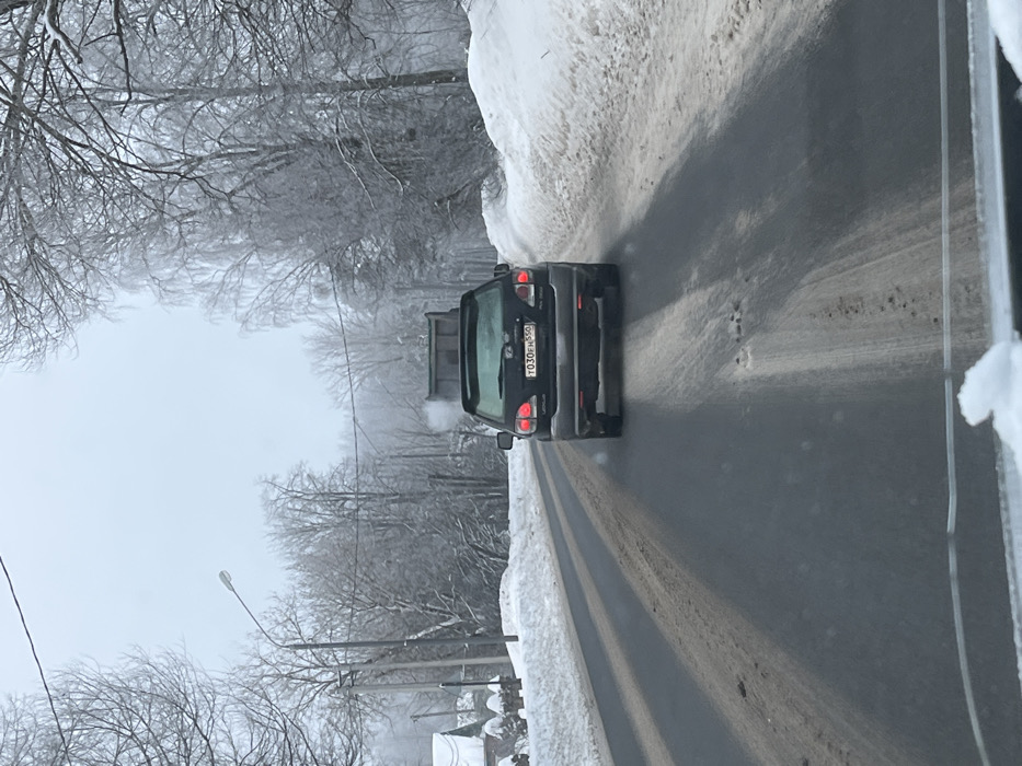 The image displays a winter road scene in Ширяево, Russia, observed from the interior of a vehicle facing forward. A dark-colored Lexus RX 300 SUV is prominently featured, viewed from the rear, positioned on a two-lane asphalt road. Its red tail lights are illuminated. The vehicle bears a Russian license plate, "T030EH 550." Ahead of the SUV, a dark-colored dump truck is visible, also traveling in the same direction, with a white plume of exhaust or steam emanating from its rear.
The road surface is partially cleared, exhibiting visible tire tracks, slush, and dirt in the lanes and central areas, along with minor depressions. Significant snowbanks line both sides of the road. The surrounding environment consists of numerous bare deciduous trees heavily coated in snow or frost. Utility poles with overhead wires are present on the left side of the road, where the faint outlines of snow-covered residential structures are also visible. A street light fixture extends from one of the utility poles. The sky is overcast and appears bright, indicating daytime conditions. No individuals are visible within the frame.