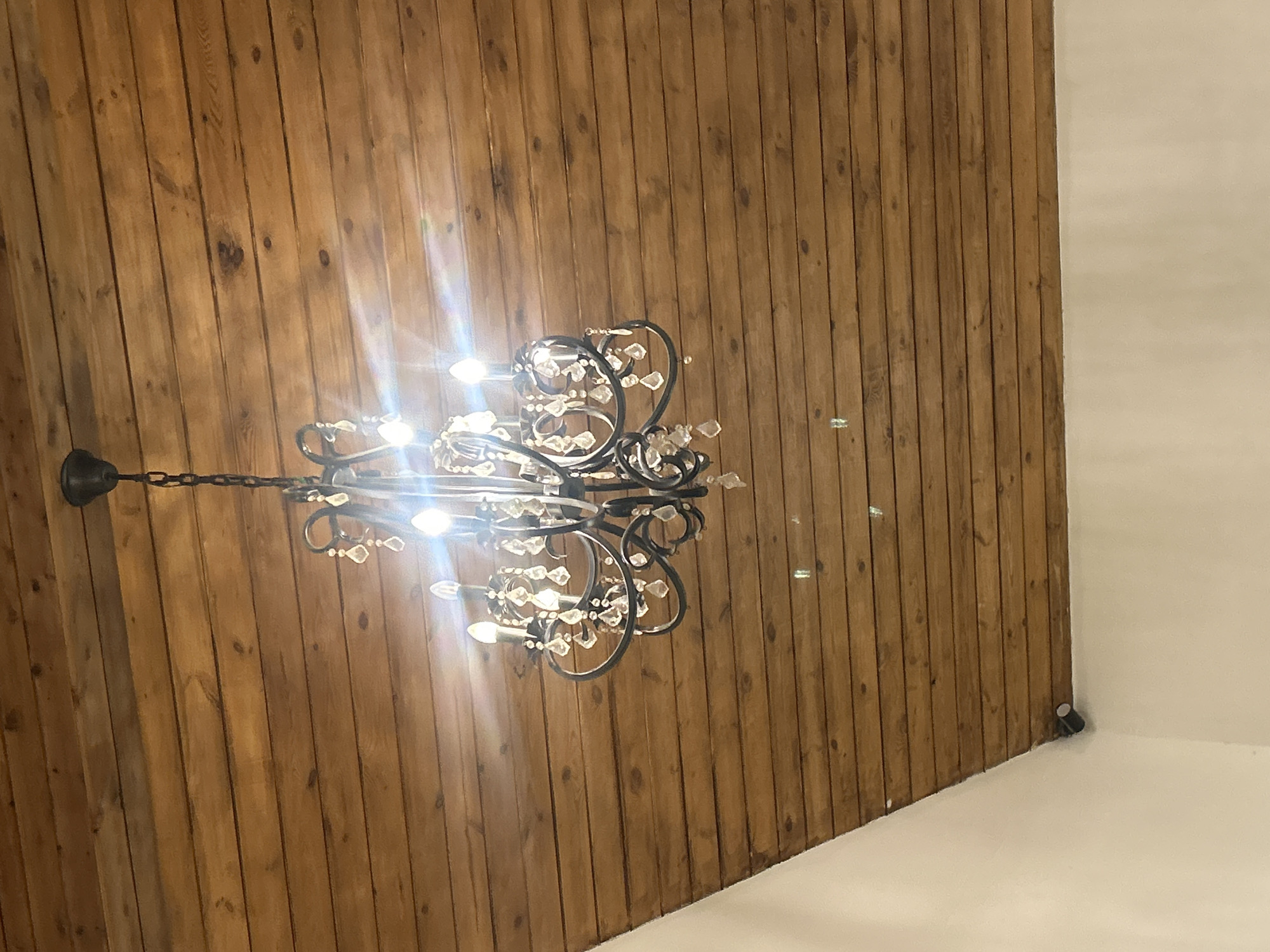 The image shows a chandelier hanging from a wooden slatted ceiling. The chandelier has a dark metal frame and is adorned with crystals. Several bulbs are illuminated, casting a bright light and creating lens flares. A portion of a white wall is visible in the lower left corner. There is a small, dark cylindrical object mounted on the wall near the corner. The setting appears to be indoors. The city of Almaty, Kazakhstan is not explicitly depicted or referenced in the image.