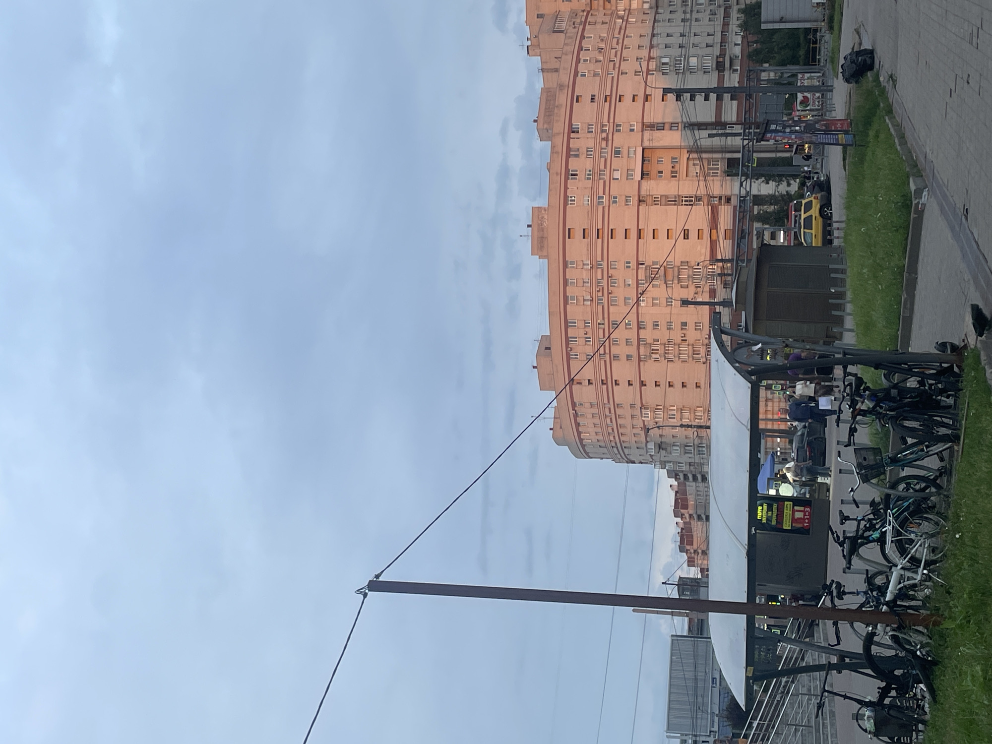 This media file depicts a street scene in Saint Petersburg, Russia, under a cloudy sky, likely in the late afternoon or early evening. The dominant subject is a large, curved, pale pink apartment building with numerous windows, stretching across the frame. In the foreground, a metal pole with wires extends upwards, creating a visual division.
To the right of the building, a paved sidewalk runs alongside a grassy strip. Several bicycles are parked near a covered structure, suggesting a public transport stop or a bike rental station. There are also a few people visible near this structure, some appearing to be waiting or interacting.
Further down the street, a yellow car is parked, and other vehicles and buildings can be seen in the background, indicating an urban environment. A black trash bag sits on the sidewalk near the edge of the frame.
There is some visible text on a sign attached to the covered structure, which appears to be in Russian. The overall mood of the image is quiet and everyday, reflecting a typical urban scene. The lighting suggests diffused daylight rather than direct sunlight.