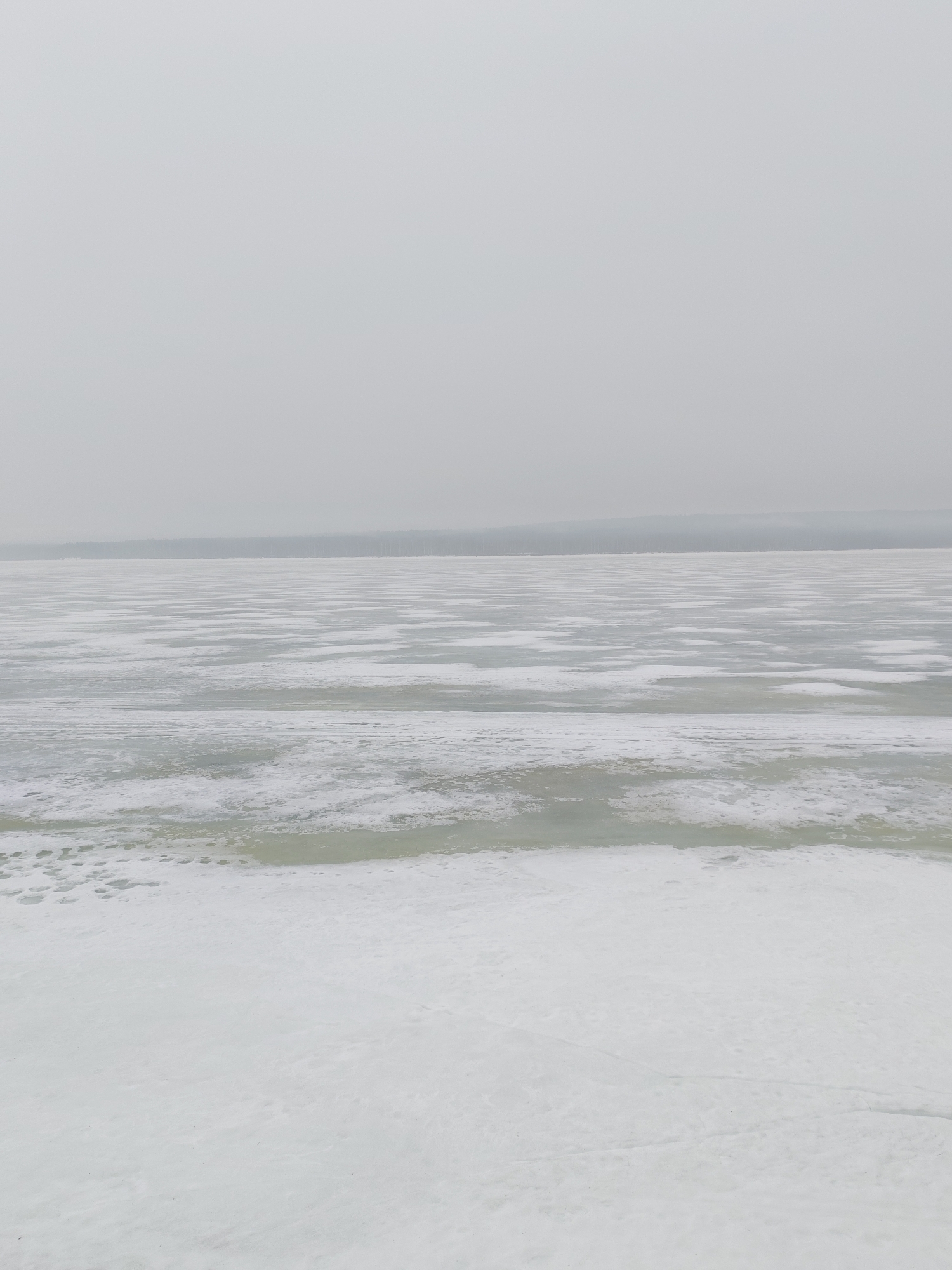 The image depicts a winter scene on the Kama River in Chaikovsky, Russia. The river is largely frozen, with ice covering the majority of the surface. Patches of open water or thinner ice are visible interspersed with the solid ice cover. The foreground consists of a snow-covered riverbank. In the background, a faint treeline can be observed on the opposite shore, and the sky is overcast, contributing to the overall gray and muted color palette. There are no people or objects immediately identifiable in the frame, focusing solely on the natural environment.