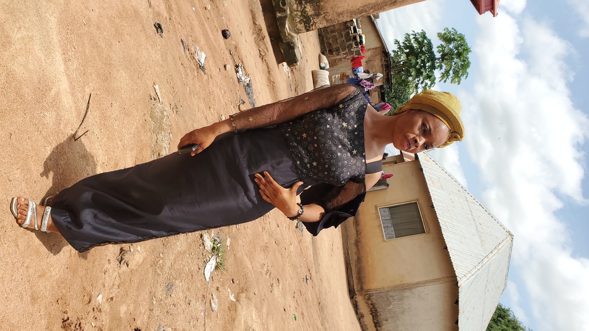 A young woman stands in an outdoor, rural setting, likely in Jalingo, Nigeria. She is wearing a black, off-the-shoulder dress with sheer sleeves adorned with gold stars. Her head is covered with a gold-colored turban. She is holding a dark-colored mobile phone in her right hand, which is resting on her hip. Her left hand is placed on her abdomen. She is wearing silver-embellished sandals. The ground is made of reddish-brown dirt, with scattered debris including plastic bags, a coconut, and twigs. In the background, there is a simple, light-colored building with a corrugated metal roof, and a smaller, similarly constructed building with a red-tiled roof. A tree with lush green foliage is visible to the left of the main building, and laundry is hanging on a clothesline in the distance. The sky is partly cloudy with white clouds against a blue background. The overall impression is that of a portrait taken in a modest, developing area.
