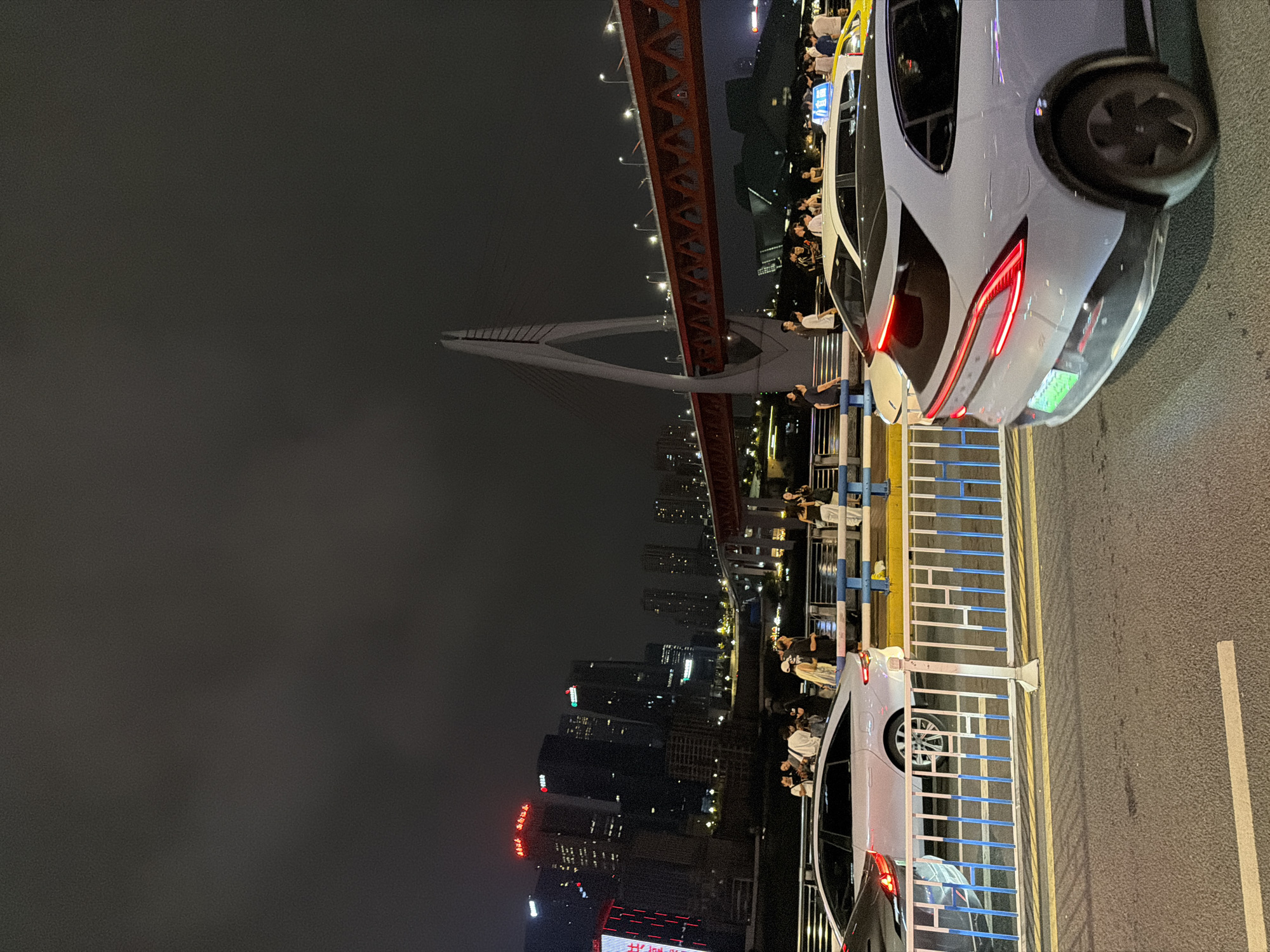 A nighttime view captures a bustling cityscape in Yuzhong District, China, characterized by a bridge spanning a body of water, illuminated by city lights and street lamps. The foreground features several vehicles, including a light-colored sedan with its taillights illuminated and a darker sedan. A white fence with blue accents separates the road from a pedestrian walkway where a group of people are gathered, some appearing to interact with their phones. In the background, the imposing architecture of skyscrapers dominates the skyline, their windows glowing with interior lights. A distinctive red bridge with intricate structural elements crosses the water, and a prominent cable-stayed bridge with a large, arching support tower is also visible. The sky is dark and overcast, contributing to the nighttime ambiance.