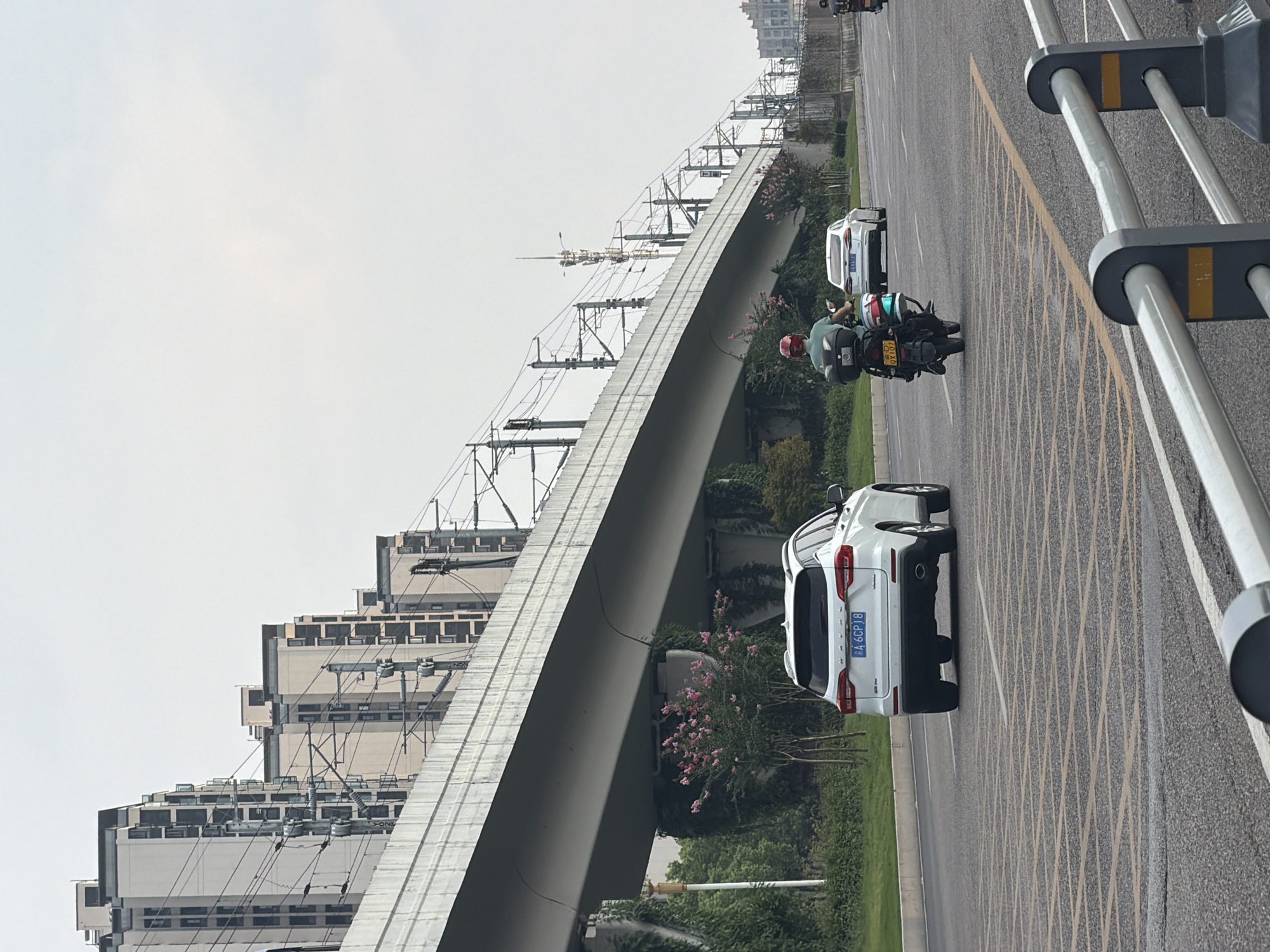 The image depicts a street scene in Jiangjin, China, under an elevated structure that appears to be a railway or highway overpass. The sky is overcast. In the foreground, a road surface marked with yellow grid lines for traffic control is visible. A white SUV with a license plate reading "A 6CP18" is driving in the middle lane. To the right of the SUV, a motorcycle with a rider wearing a red helmet is also in motion. The motorcycle has a license plate reading "QX107". Further in the background, a white sedan is visible. Along the side of the road, there are trees with pink flowers, and ivy is growing on the support structures of the overpass. In the distance, a series of tall, multi-story buildings with a consistent architectural style line the horizon, suggesting an urban or suburban residential area. Electrical poles and overhead wires associated with the overpass are prominent.