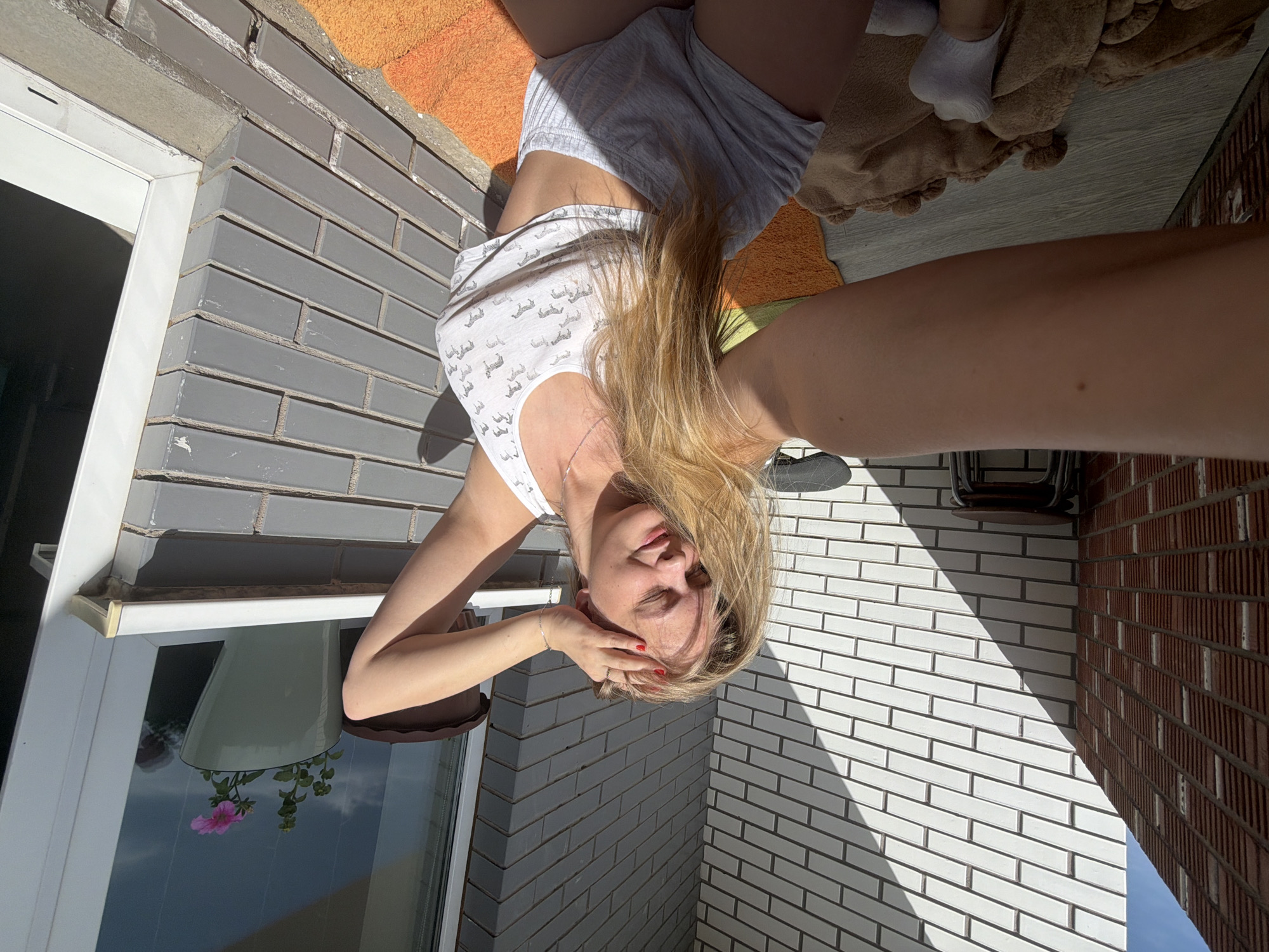A woman is lying on a balcony, facing the camera which is positioned above her. She has blonde hair and is wearing a white tank top and grey shorts. Her hands are near her face. A window with a flower pot is to her left. The balcony has grey and white brick walls, and a section of brick is a darker red. Towels are to her right. The location may be a balcony in Izhevsk, Russia, as per the user's request.