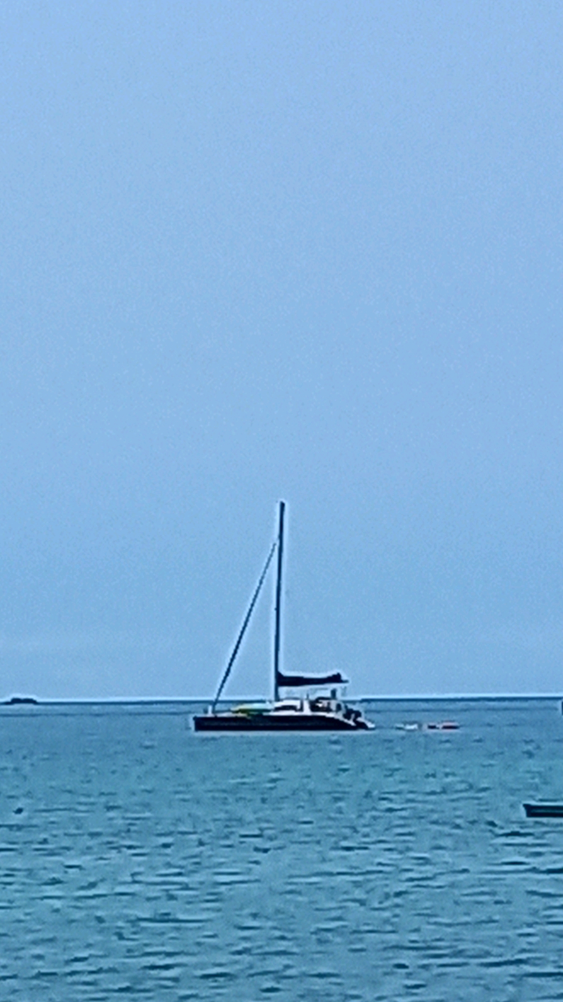A catamaran sailboat is anchored in the clear blue waters off the coast of Ko Pha-ngan District, Thailand. The sky is a pale blue and the water is calm with small ripples. The catamaran has a white hull and two masts, with one sail partially furled. There are a few people visible on the deck of the boat. In the distance, a small island can be seen. The scene appears tranquil and serene, suggesting a peaceful day at sea.