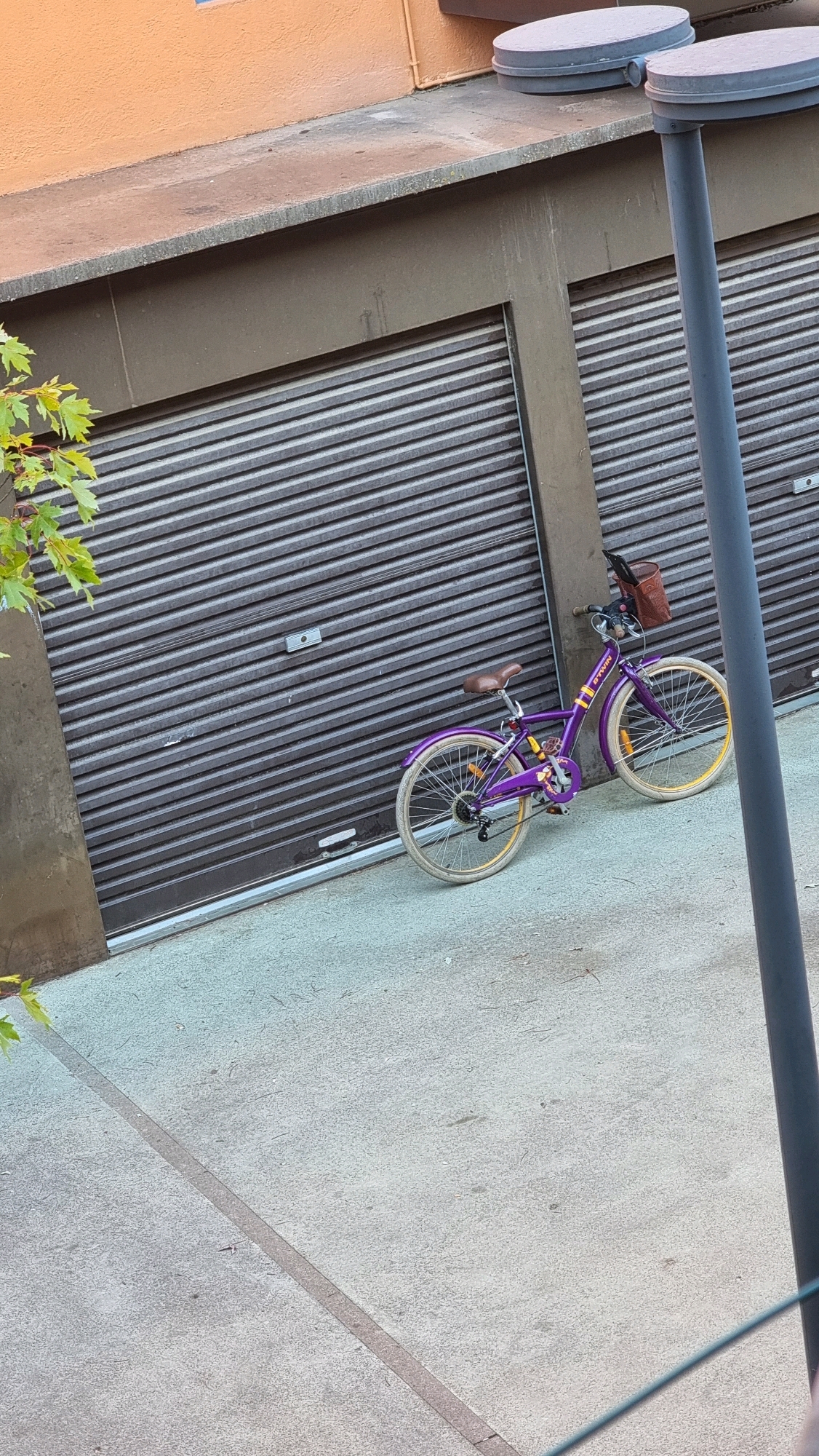 A purple bicycle with a brown basket is parked in front of a dark gray roller door. The bicycle has a brown leather seat and handlebars. The wheels have yellow rims. To the left of the bicycle, a few green leaves from a tree are visible, with branches extending out of frame. Behind the bicycle and to its right, a dark gray pole stands vertically. The roller door is part of a building with an orange wall above it. The ground in front of the roller door is a light gray concrete surface, with a diagonal seam running across it. The scene appears to be an outdoor urban environment, possibly a courtyard or alleyway, in Sant Joan Les Fonts, Spain.