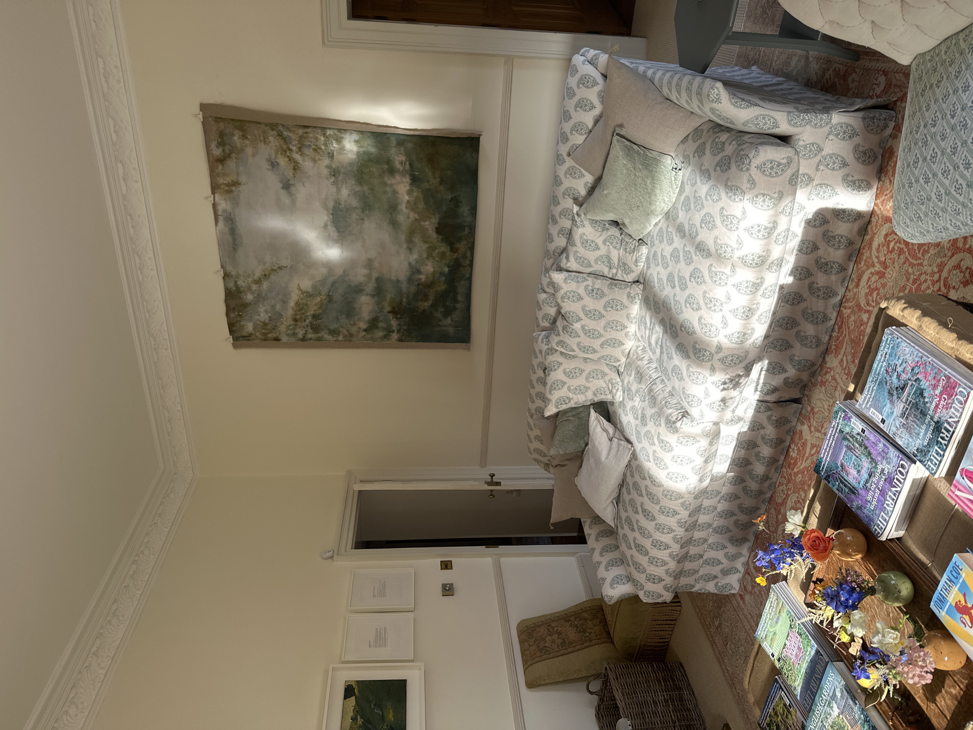 The image shows an interior room with an eclectic and somewhat rustic style. The walls are painted a light cream color, and there's decorative molding along the ceiling. A large landscape painting hangs on the left wall, depicting a somewhat abstract scene with greens and grays, possibly representing trees or a forest.
A patterned sofa is situated to the right, covered in a white fabric with a subtle green or blue paisley design. Several pillows in varying colors and textures are scattered on the sofa, including beige and light green.
In the foreground, there's a coffee table adorned with stacks of magazines, specifically "Country Life," and flower arrangements in small vases. The flowers include blue and white varieties.
Behind the sofa, a glimpse of another room is visible through an open doorway. There are framed pictures or artwork on the wall near the doorway. A textured cushion or rug is on the floor next to the sofa, along with a wicker basket.
The room appears to be well-lit, possibly by natural light coming from a window or doorway not visible in the image. The location is unknown.