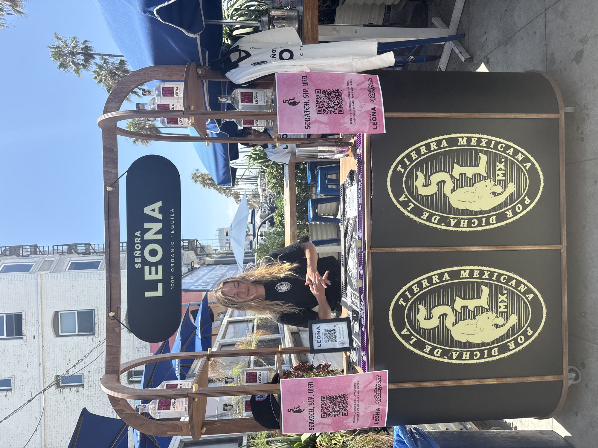 A blonde woman with long, wavy hair is standing behind a bar and presenting a scratch-off ticket with a QR code. She is wearing a black t-shirt with a circular logo. The bar is dark brown with gold trim, and features a logo with the text "SEÑORA LEONA MEXICANA" and a jaguar. There are bottles of "SEÑORA LEONA 100% ORGANIC TEQUILA" on shelves above the bar, and promotional signs with QR codes. A white shirt with a black logo is hanging on a rack to the right of the bar. The scene appears to be outdoors, possibly on a boardwalk in Santa Monica, California, given the presence of palm trees and the overall beachy atmosphere. The sky is clear and blue, suggesting a sunny day.