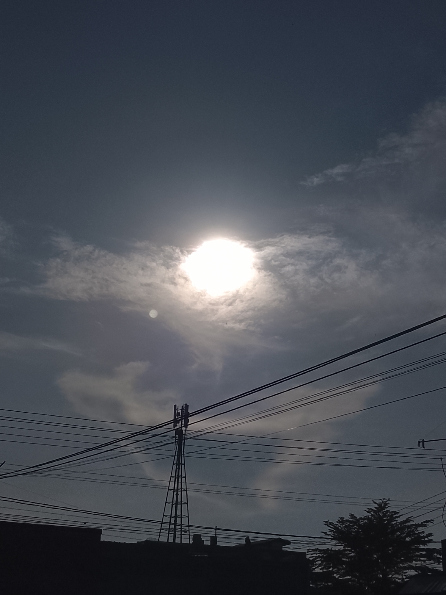 A silhouetted cell tower stands tall against a cloudy sky, with the bright sun partially obscured by the clouds, creating a hazy glow. Several power lines are strung across the frame, adding a utilitarian element to the scene. In the foreground, the dark silhouettes of buildings and a tree suggest an urban or suburban setting in Lhokseumawe, Indonesia. The overall mood is somewhat somber due to the overcast sky and muted colors.